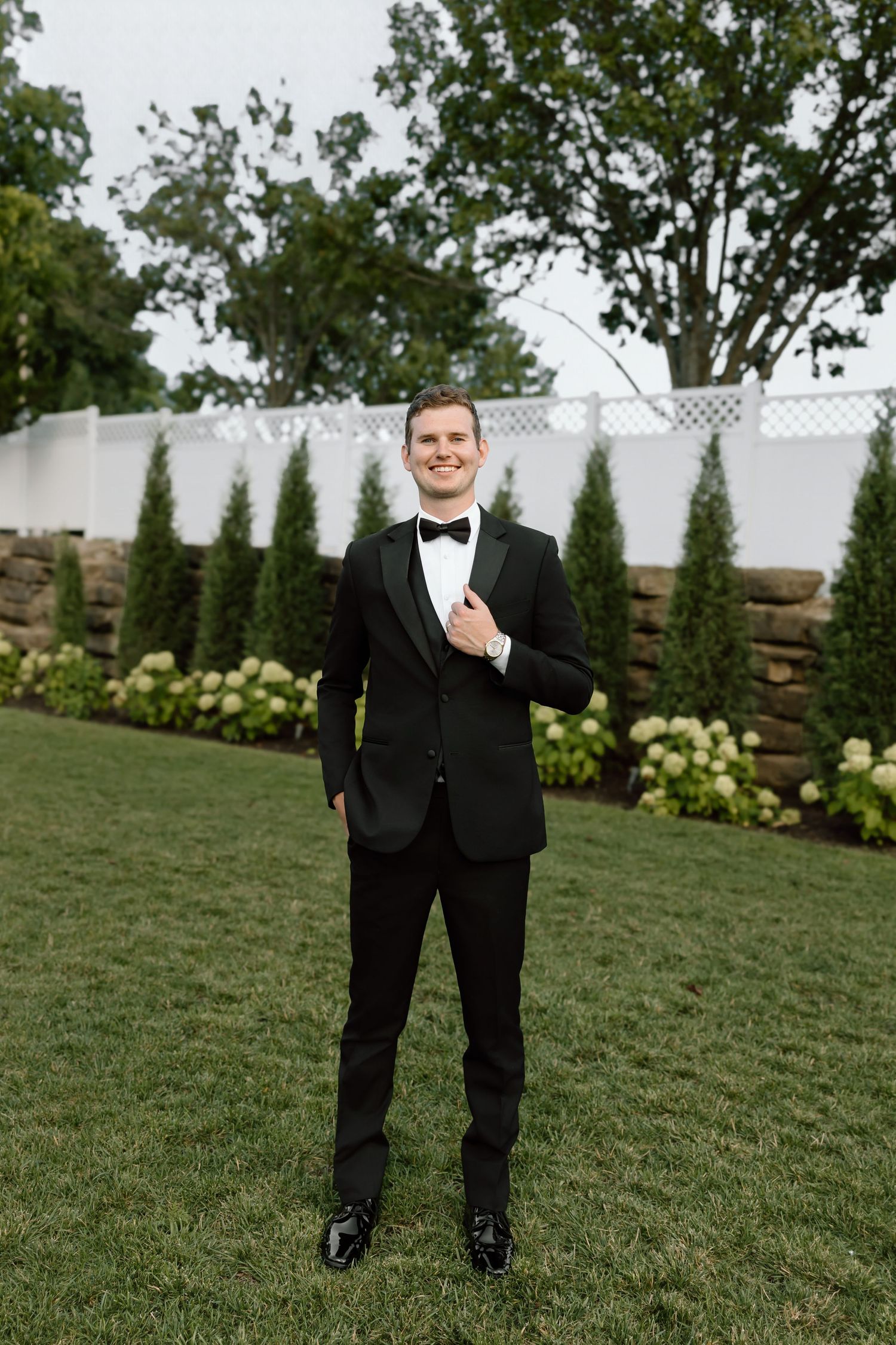 A smiling groom in a classic black tuxedo with bow tie stands on a manicured lawn surrounded by lush greenery.