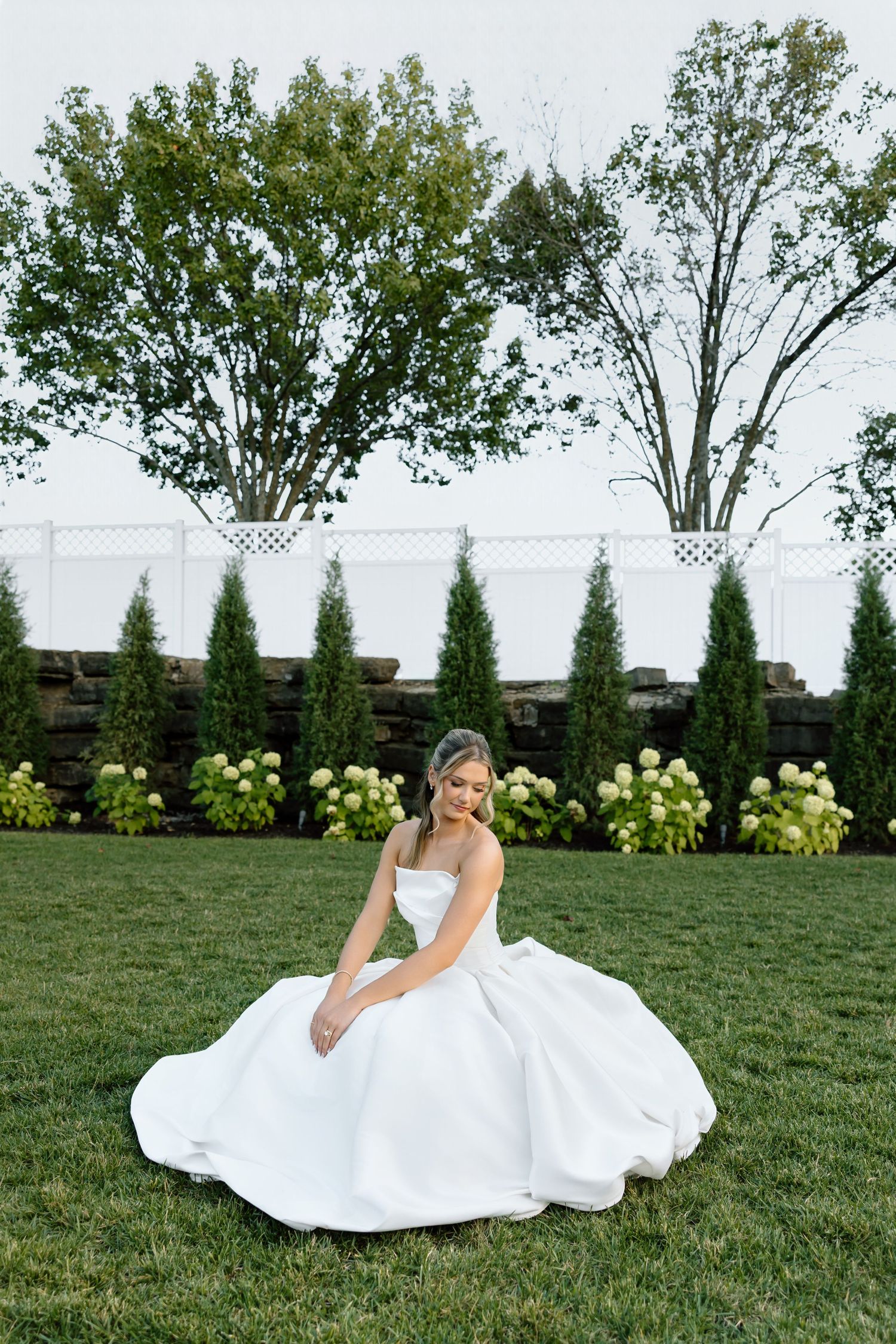Bride in a white ball gown sitting on green grass surrounded by evergreen trees and hydrangeas at an outdoor wedding venue.