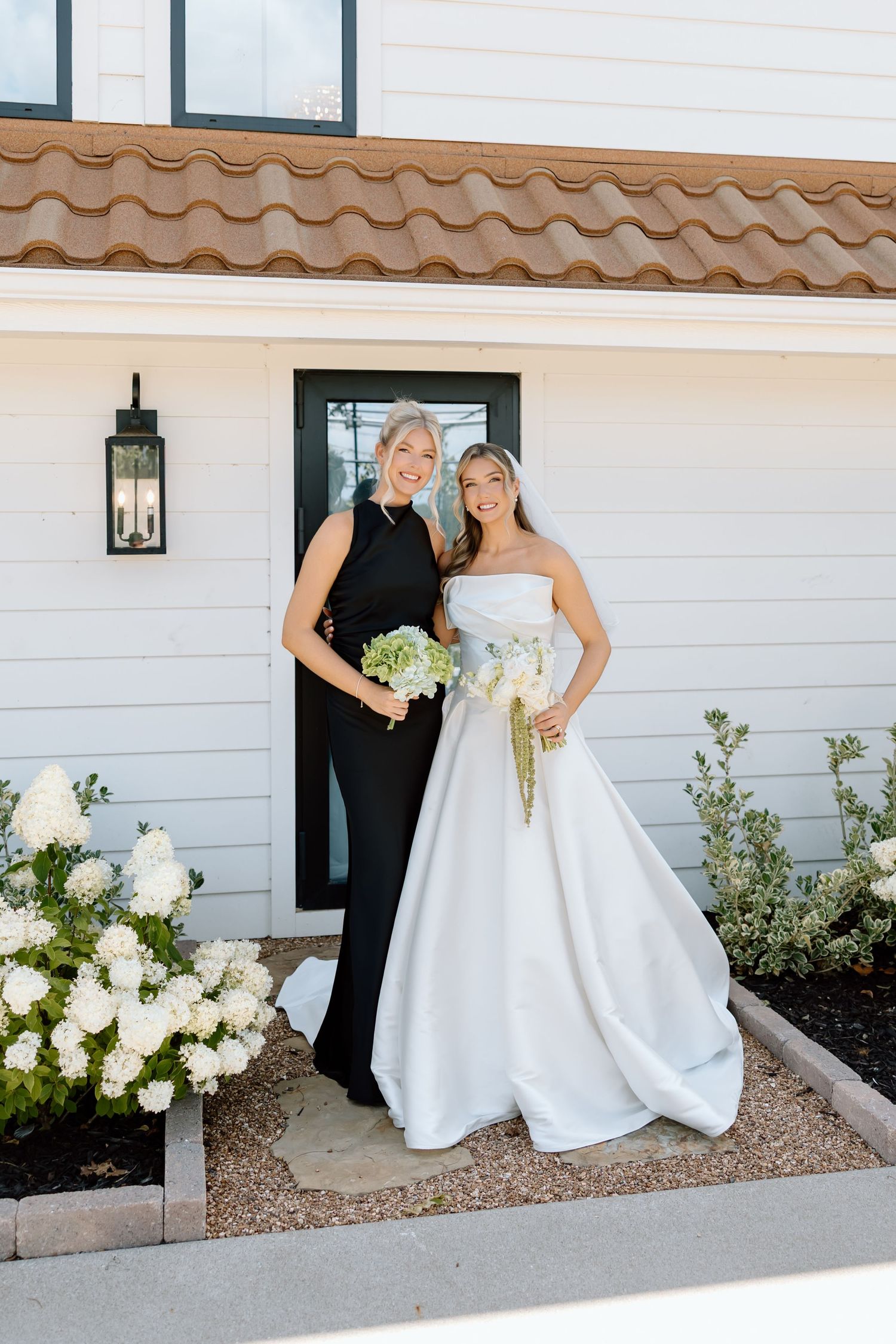 Bride in strapless white ball gown holding bouquet poses with woman in black dress outside white farmhouse.