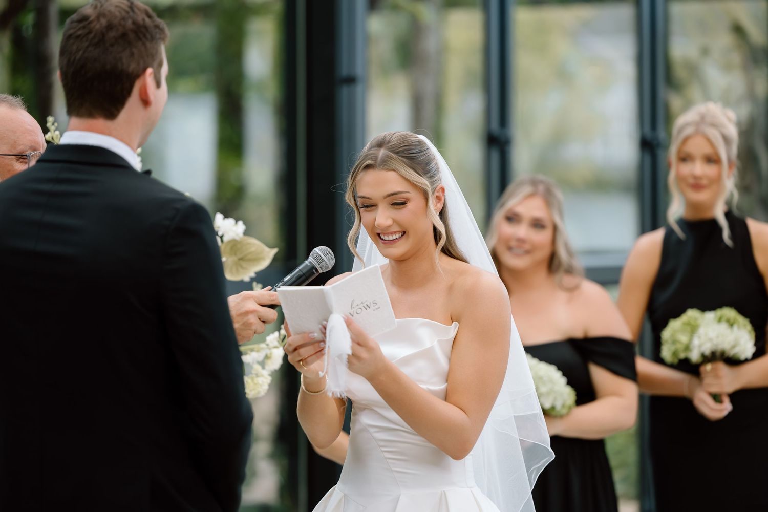 Smiling bride in white gown reads wedding vows at altar with bridesmaids in black dresses standing behind her.