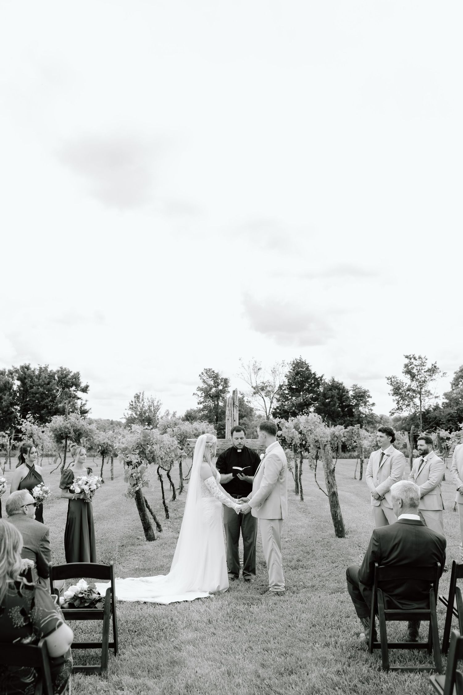 Black and white outdoor wedding ceremony in a vineyard with bride and groom holding hands before an officiant.