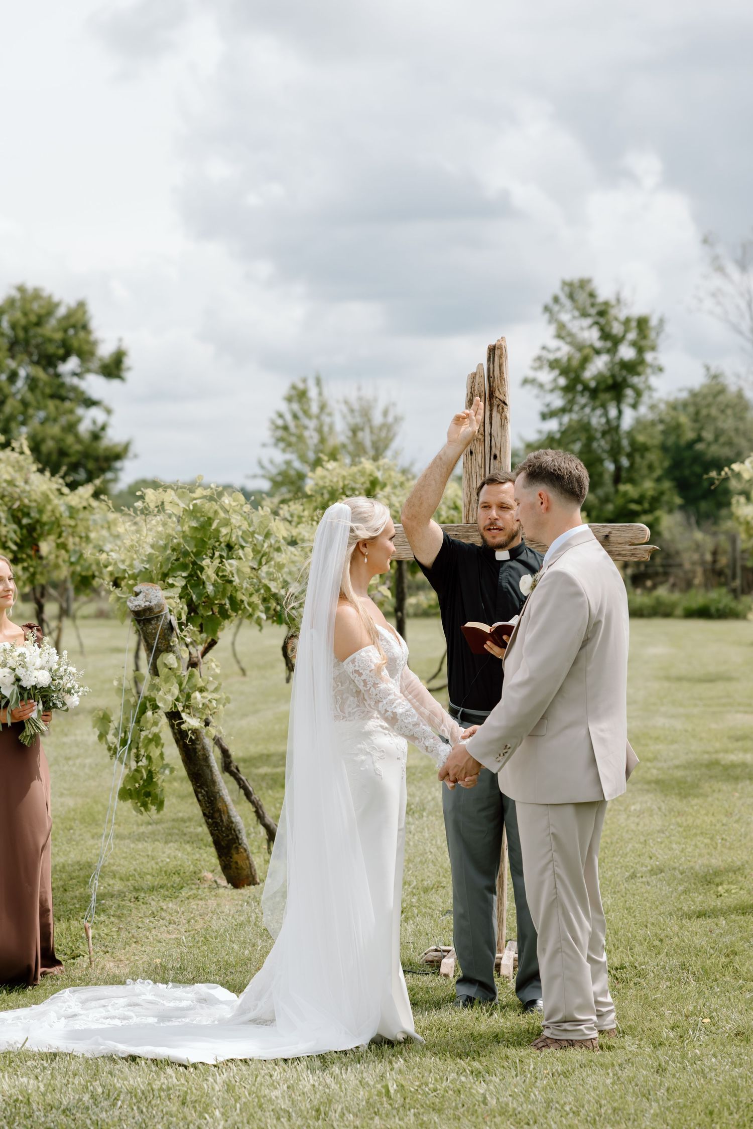 Outdoor wedding ceremony with bride in white gown, groom in tan suit, and officiant raising hand under rustic wooden arch.