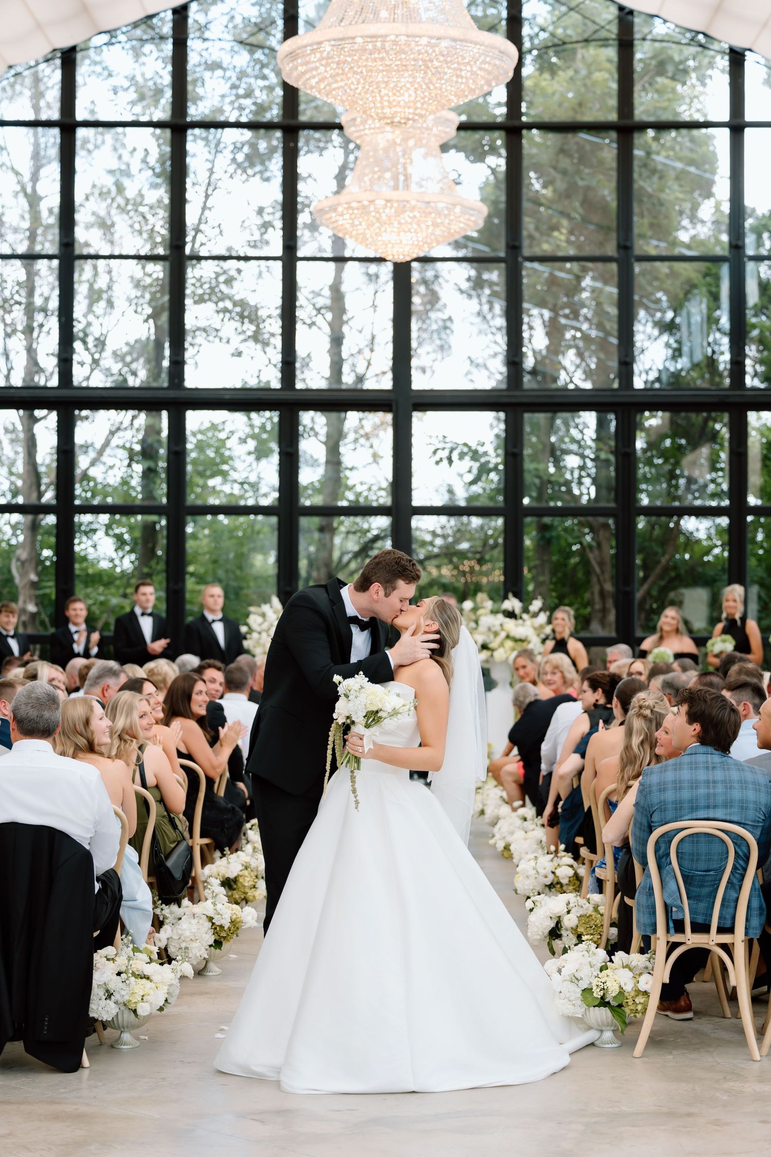 Bride and groom share first kiss at elegant indoor wedding ceremony with chandelier and floor-to-ceiling windows.