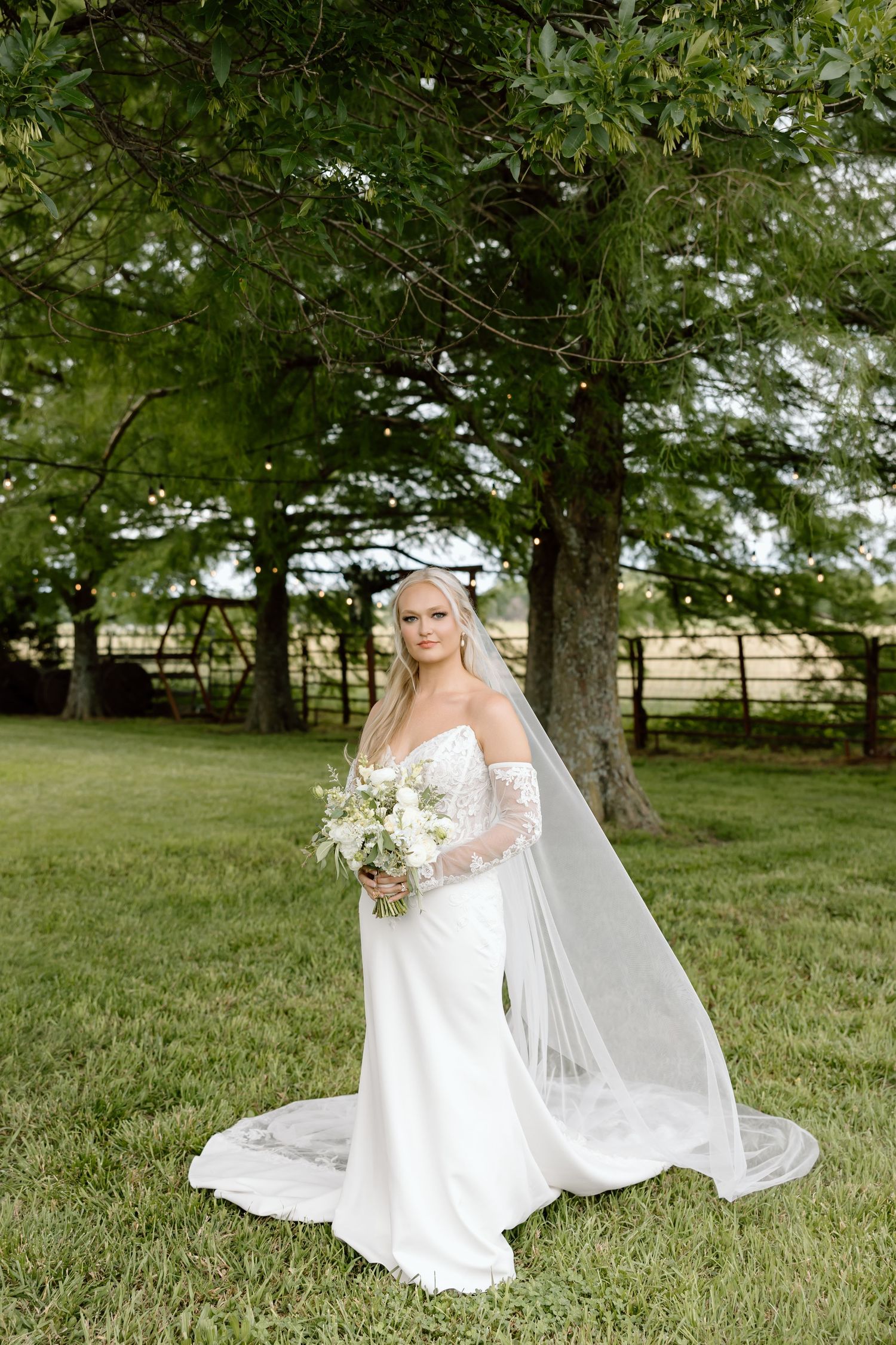 Bride in off-shoulder white gown and veil holds bouquet outdoors near large tree on green lawn.