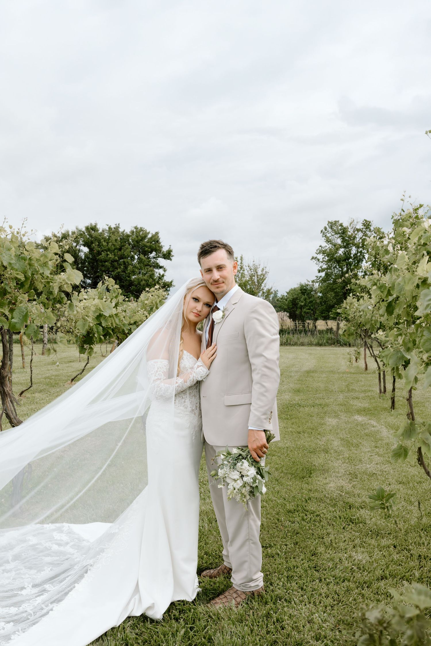 Bride and groom pose in vineyard, her veil flowing in wind, wearing white gown and beige suit, holding white bouquet.