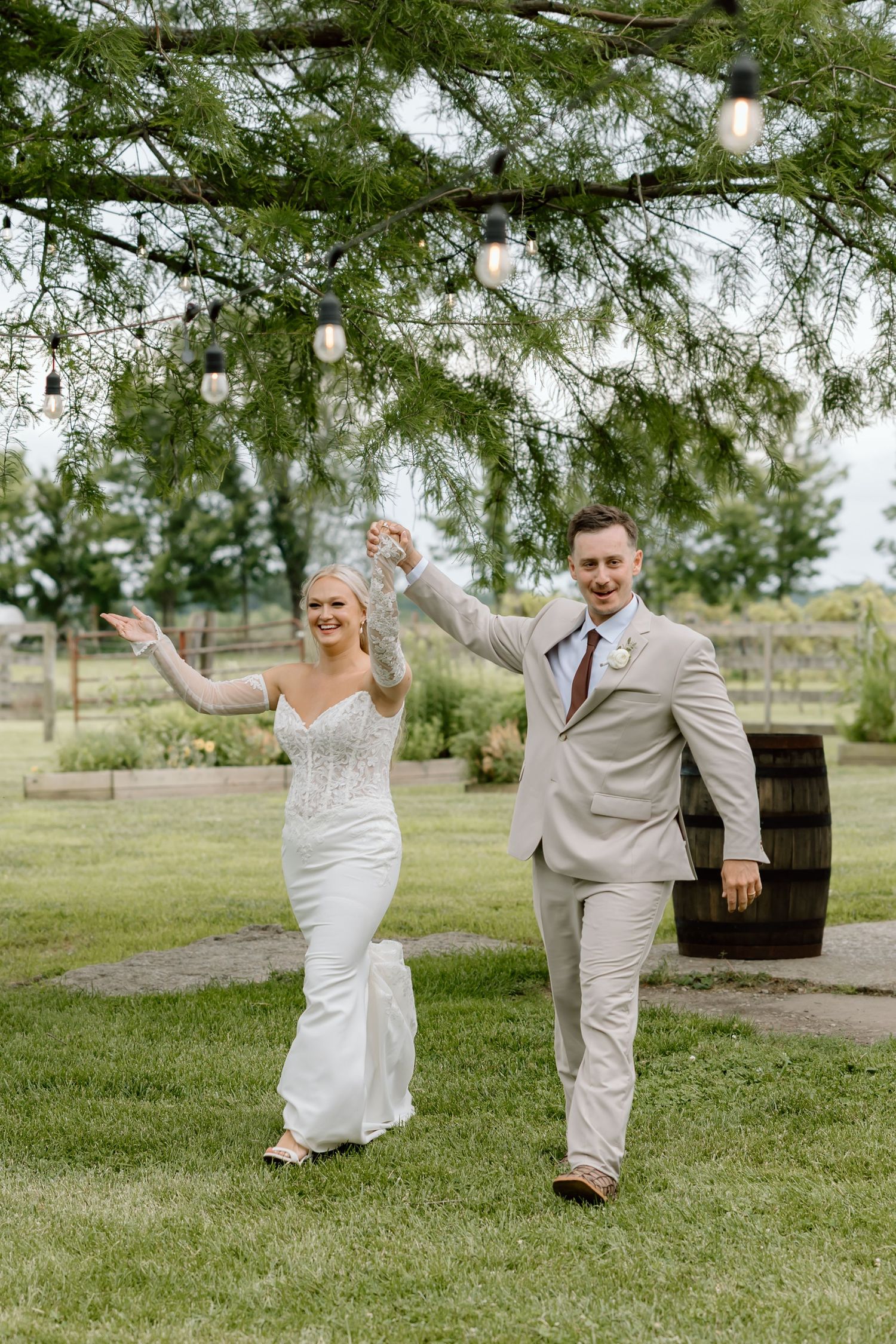 A joyful bride and groom walk hand-in-hand under string lights on a lush green lawn at an outdoor wedding venue.