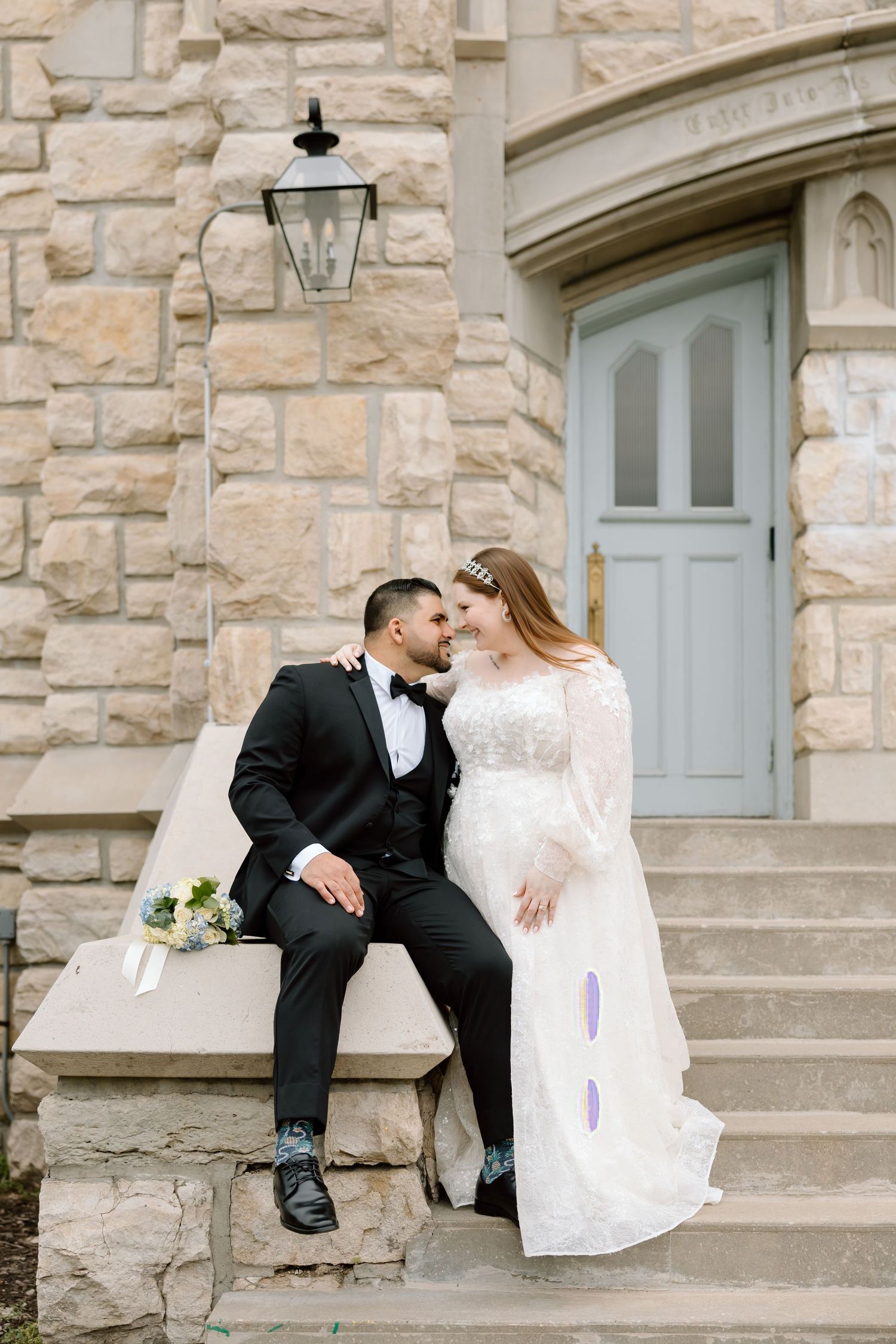 A bride and groom share a romantic moment on stone steps outside a historic building with a blue door.