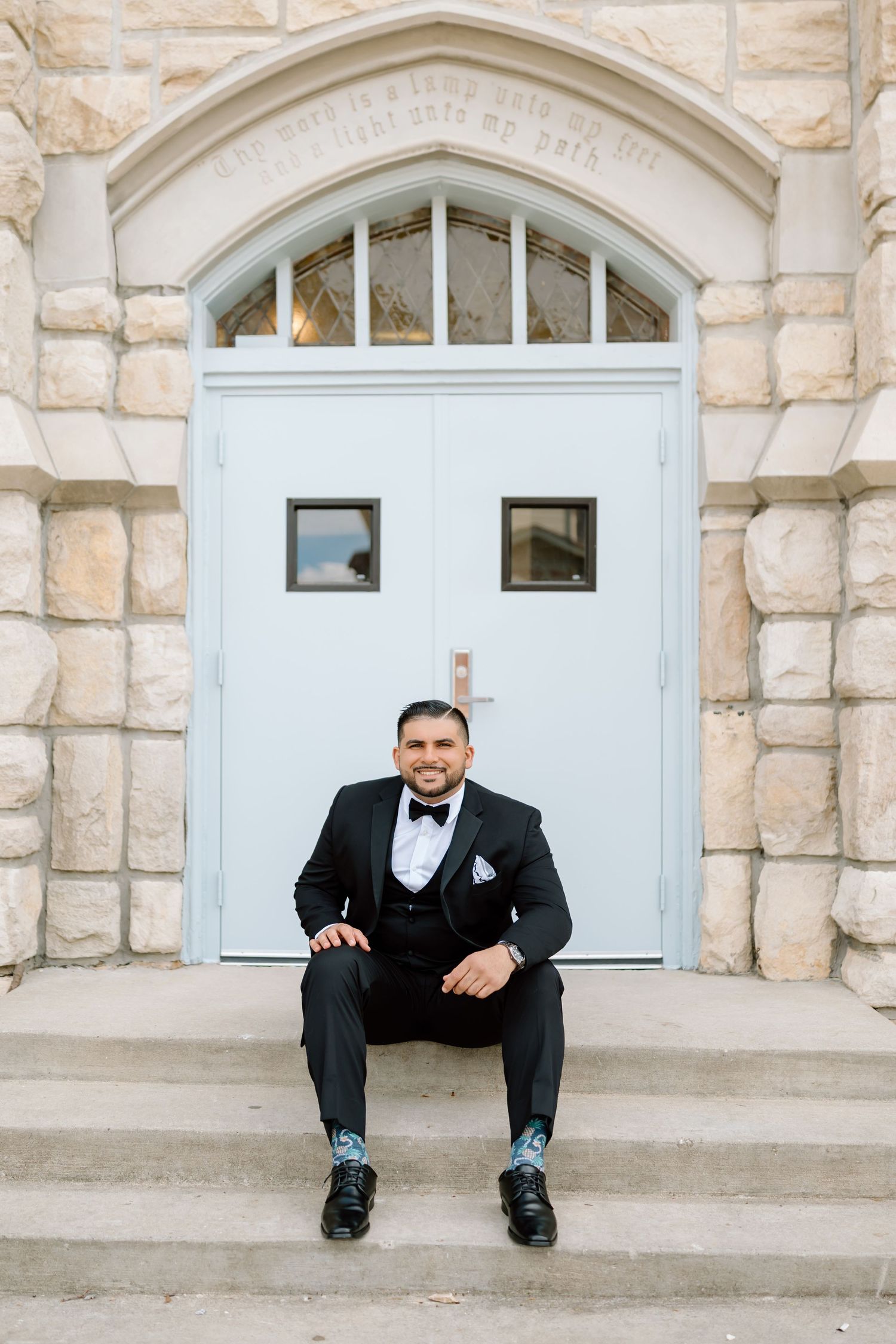 Groom in black tuxedo with bow tie sits smiling on church steps, showcasing patterned socks and pocket square.