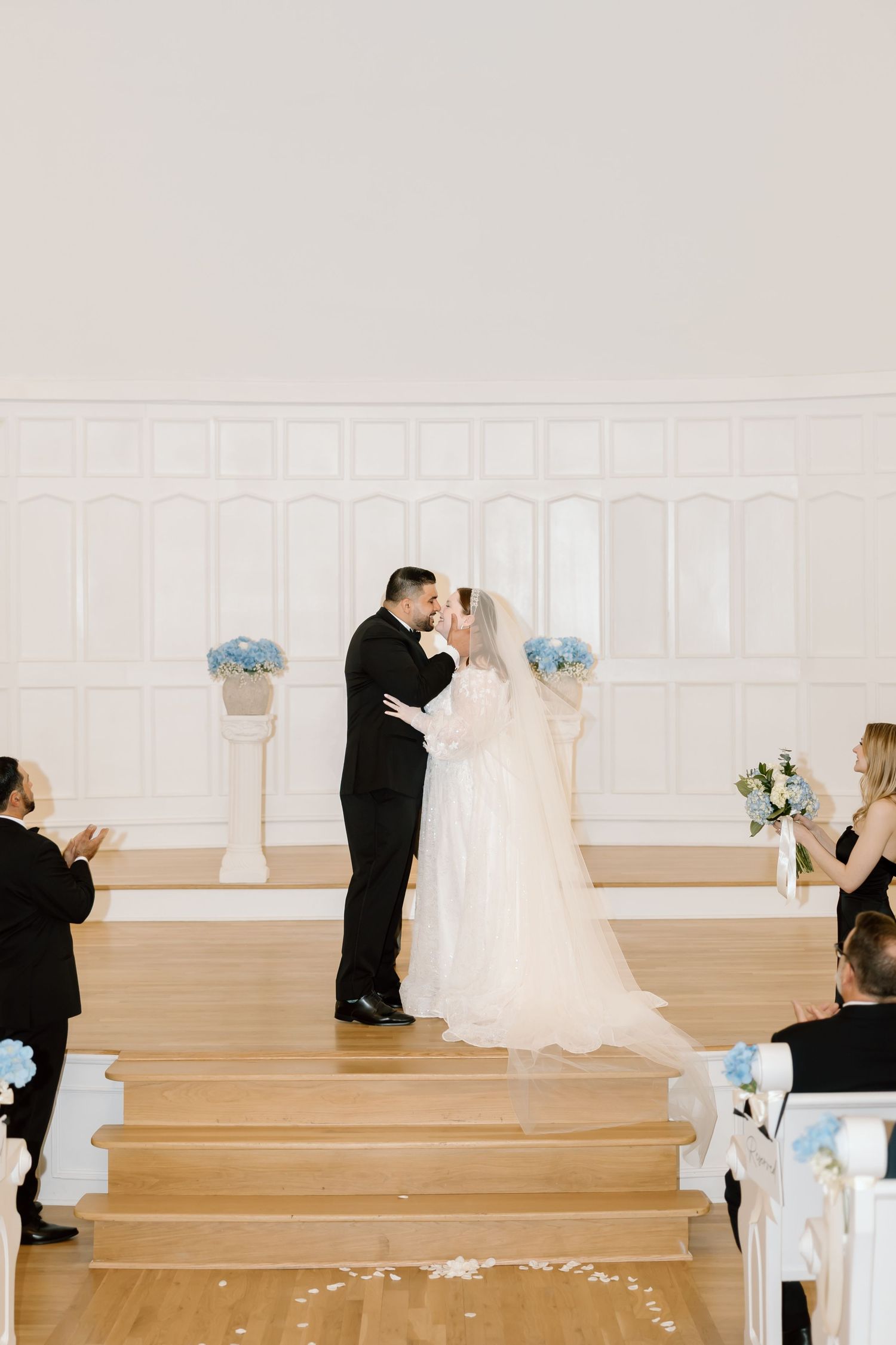 Bride and groom share first kiss at elegant white church wedding ceremony with blue floral arrangements.