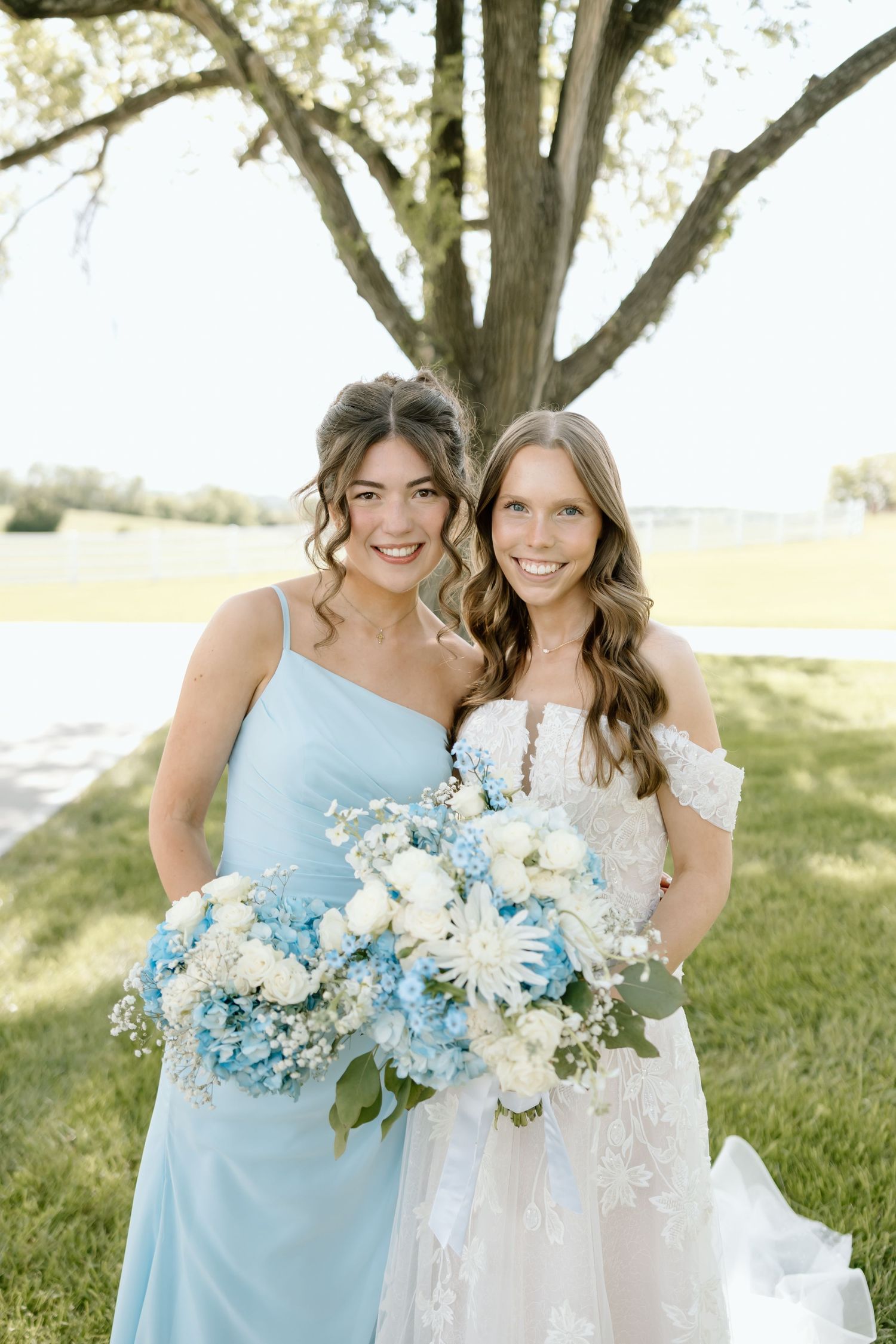 Bride in white lace gown and bridesmaid in light blue dress holding blue and white floral bouquets outdoors.