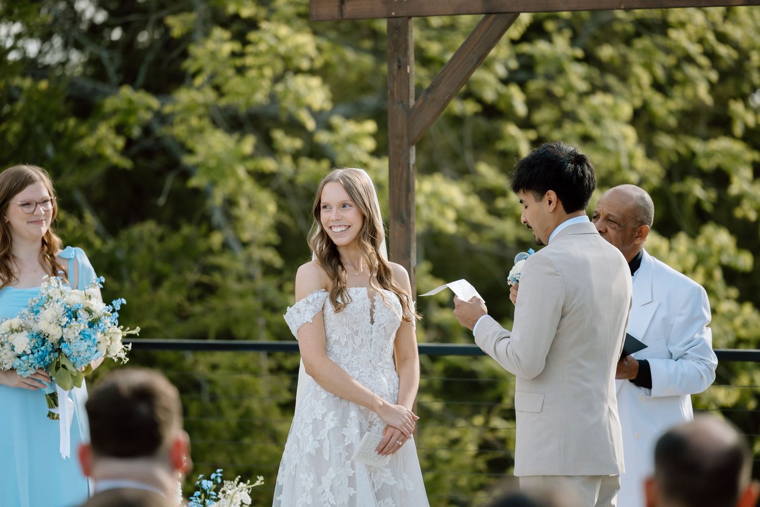 Bride smiling at groom during outdoor wedding ceremony with bridesmaid holding blue bouquet.
