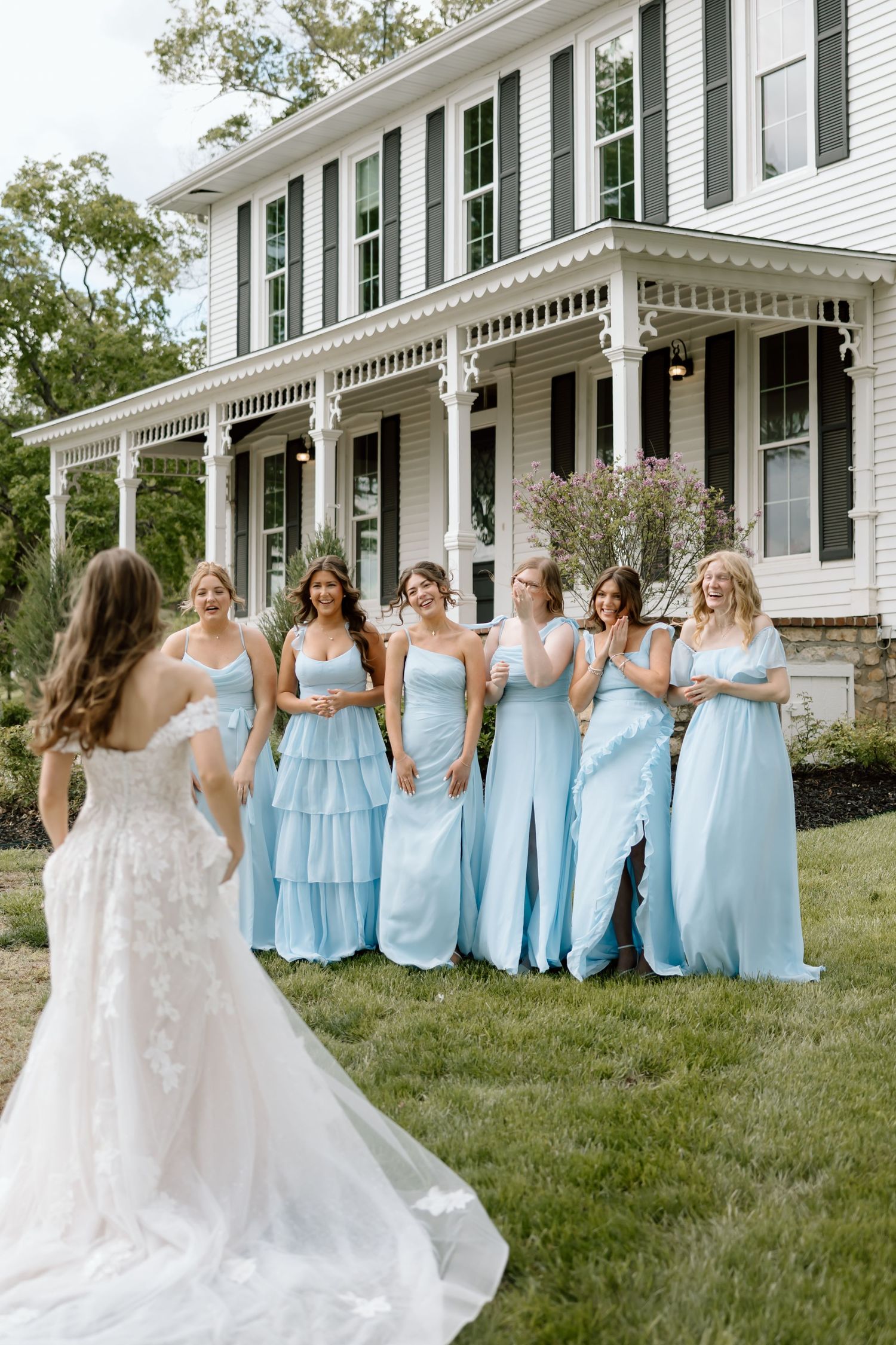 Bride in lace gown reveals herself to six bridesmaids in light blue dresses outside a white Victorian home.