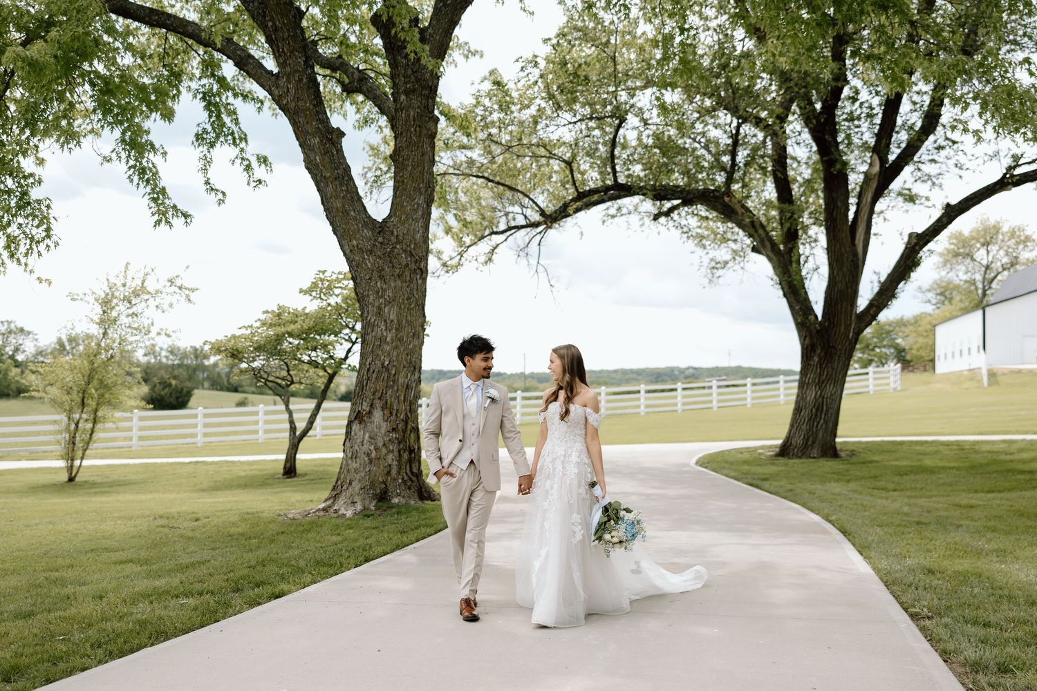 A bride and groom walk hand-in-hand along a curved path surrounded by large trees and a white fence.