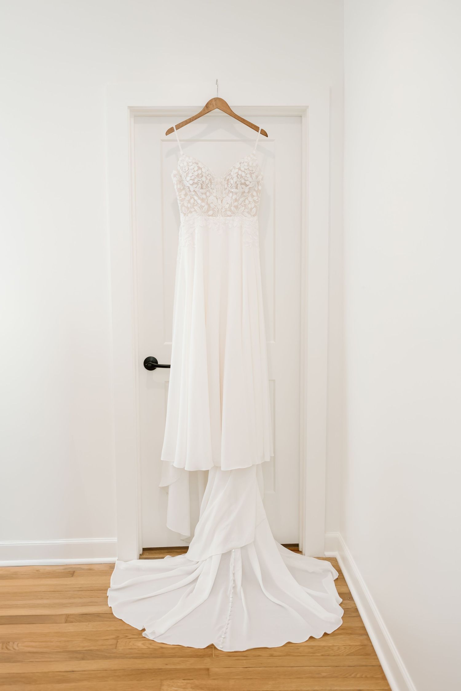 A white wedding dress with lace bodice and flowing train hangs elegantly on a wooden hanger against a white wall.