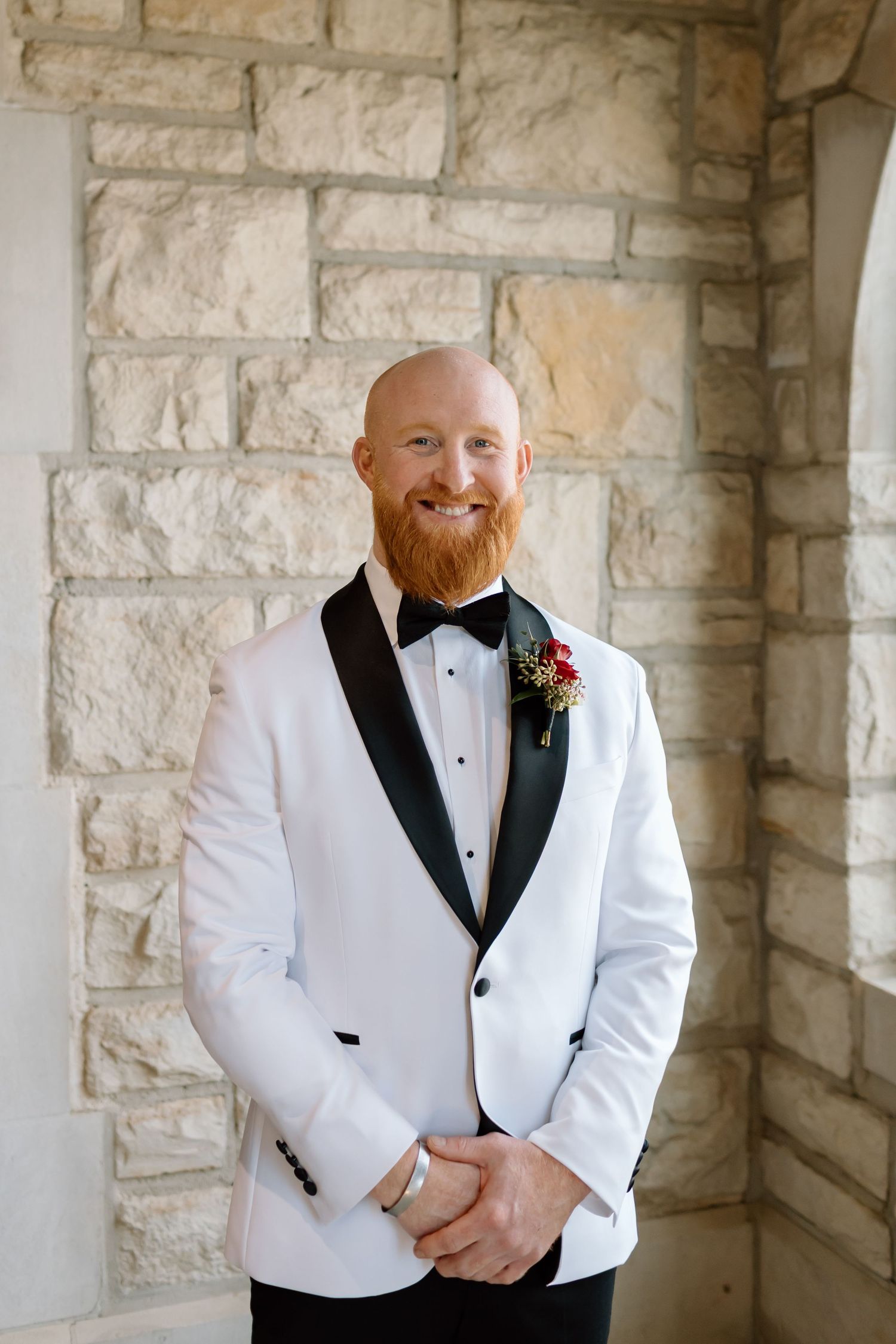 A groom in a white tuxedo jacket with black lapels poses in an elegant stone archway setting.