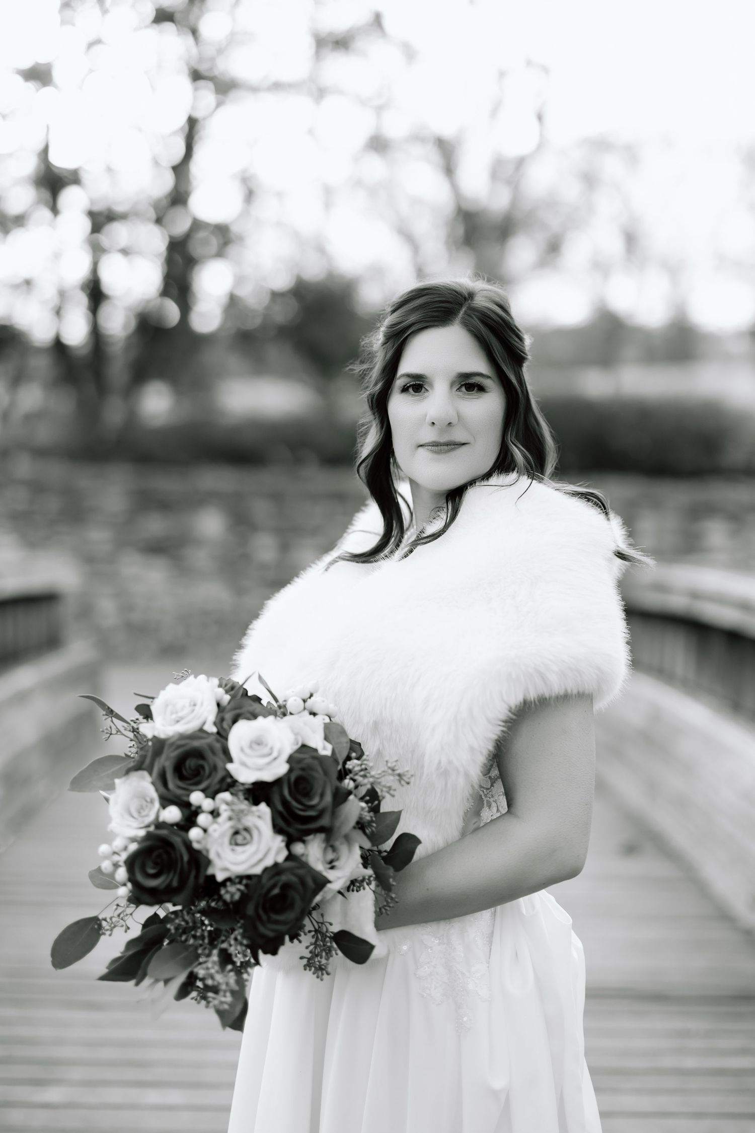 Black and white bridal portrait series showing winter wedding details with fur wrap and dark floral bouquet on wooden bridge.