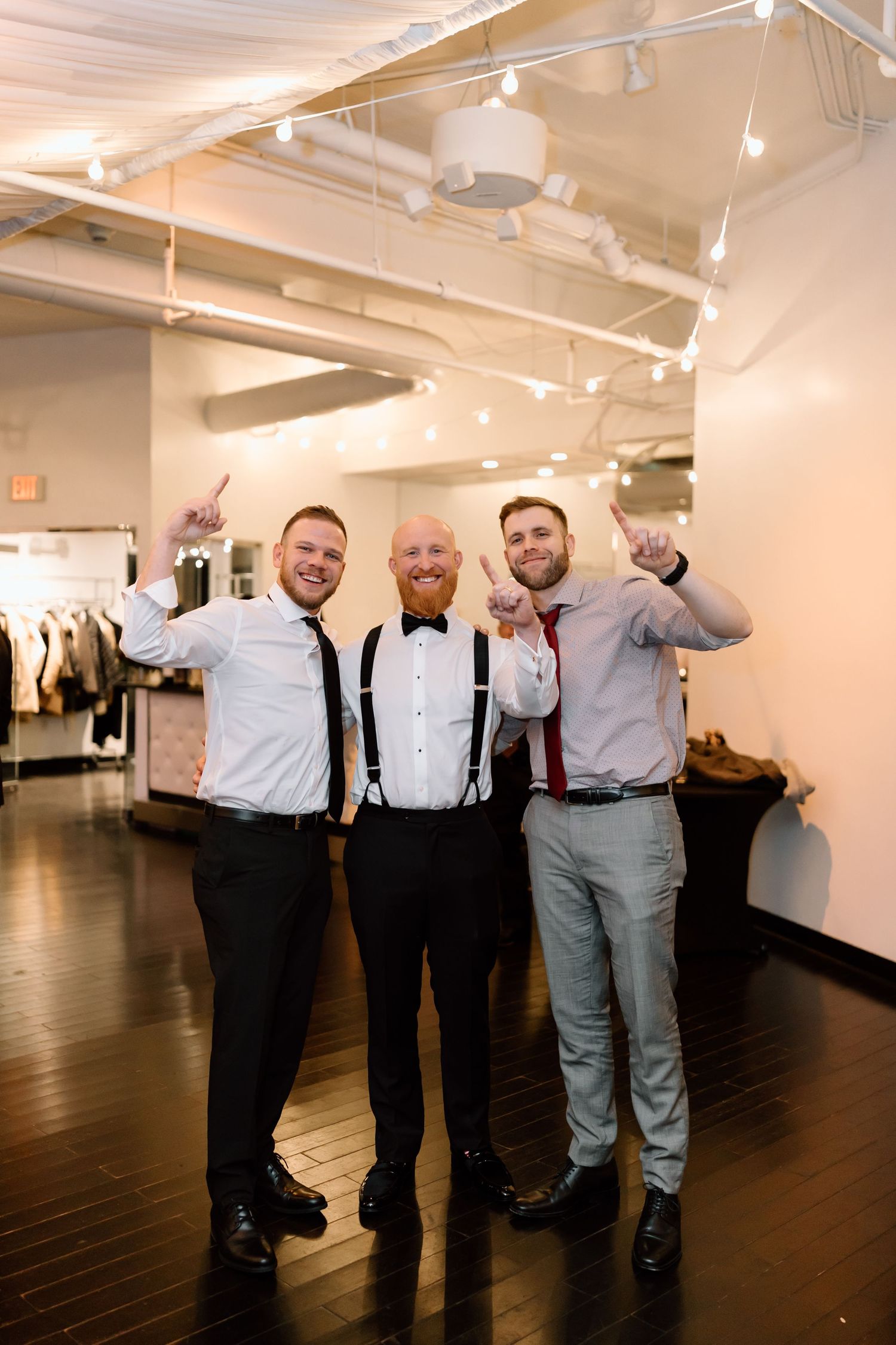 A group of groomsmen in dress shirts and suspenders pose playfully in a spacious venue with dark wood floors.