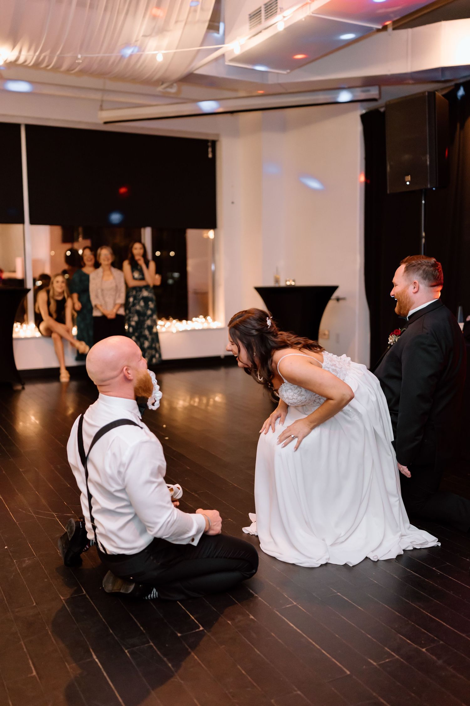 Wedding party performs fun dance moves on dark hardwood floor during reception celebration.