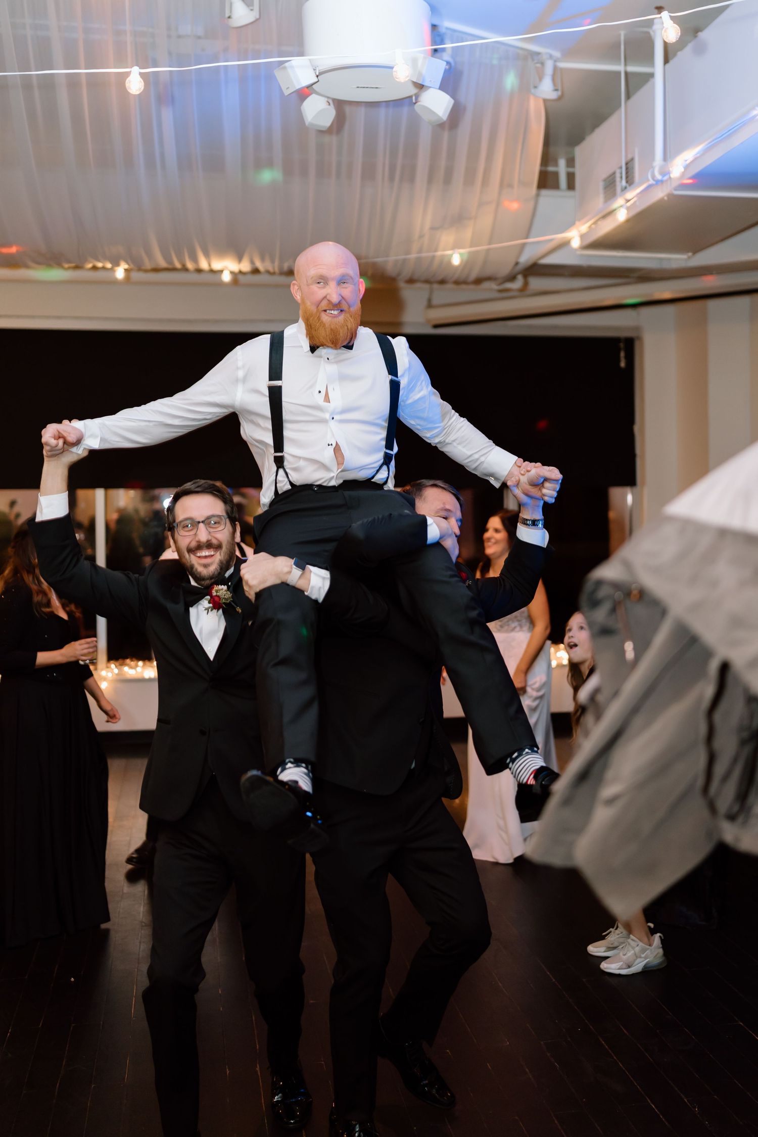 Wedding guests lift and carry someone wearing suspenders on the dance floor during a celebration.