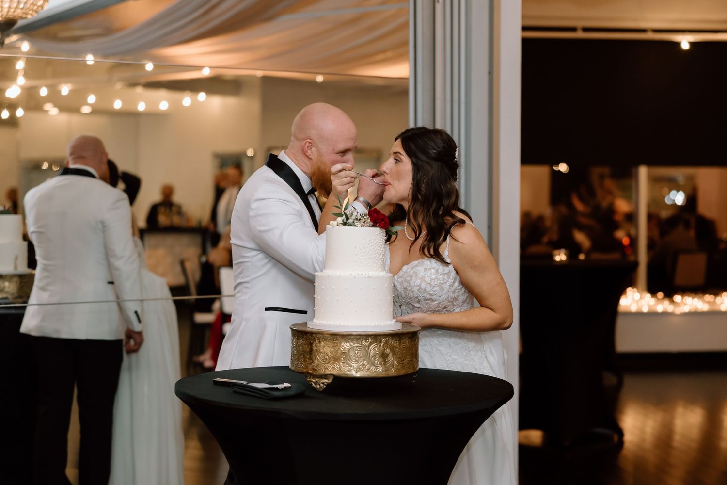 Newlyweds celebrate their first cake cutting moment together at an indoor wedding reception with string lights.