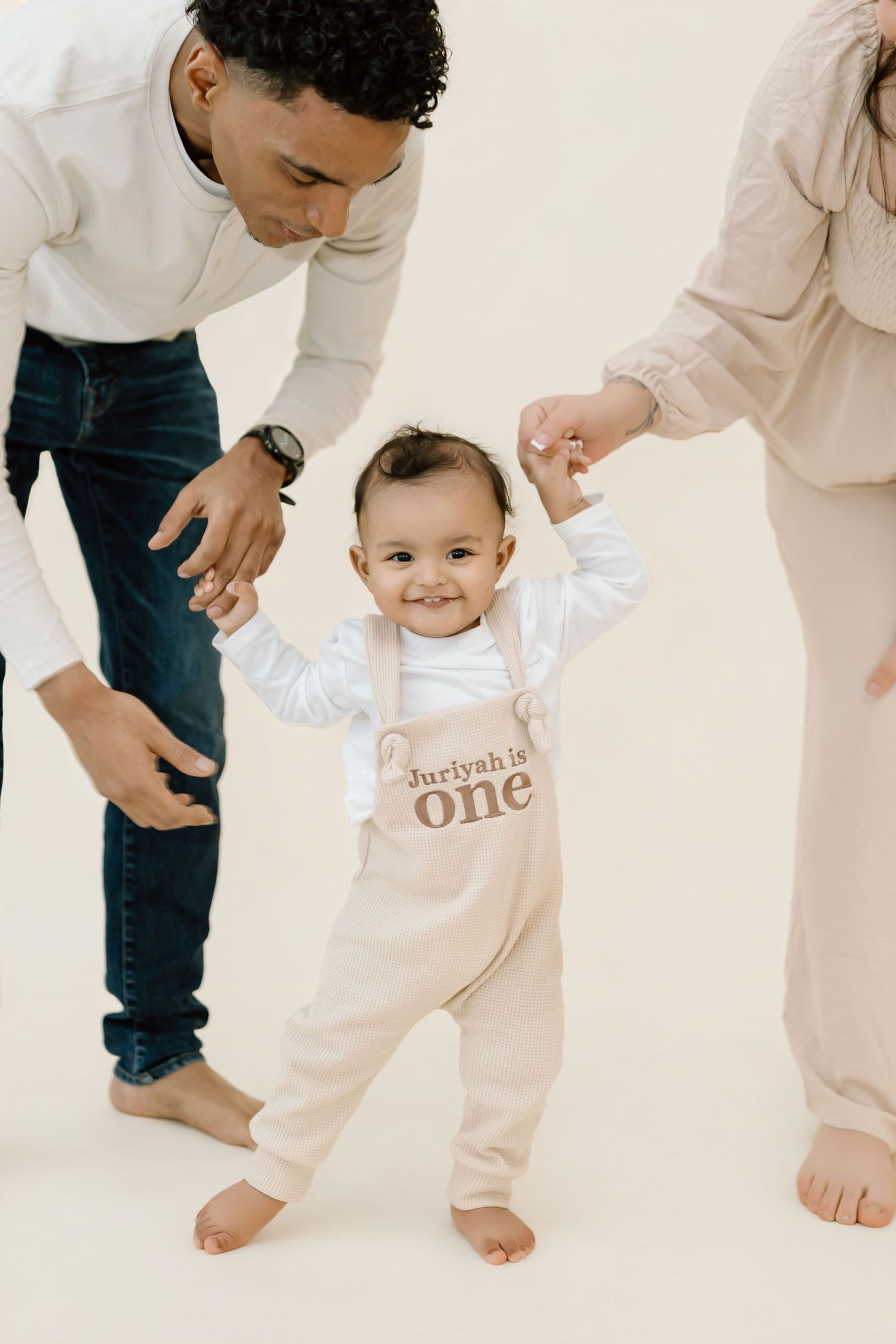 Baby in white outfit crawls on floor while adults in white and jeans look on during first birthday photoshoot.