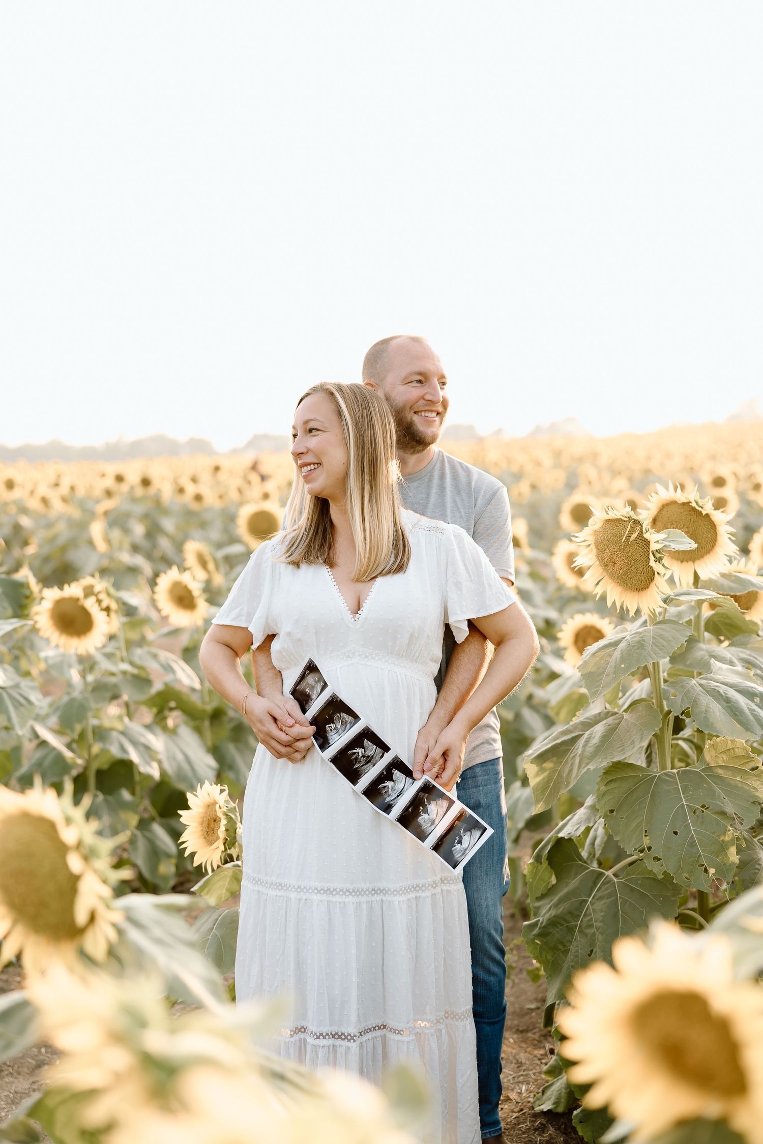 A couple stands together in a golden sunflower field during sunset holding letter signs.