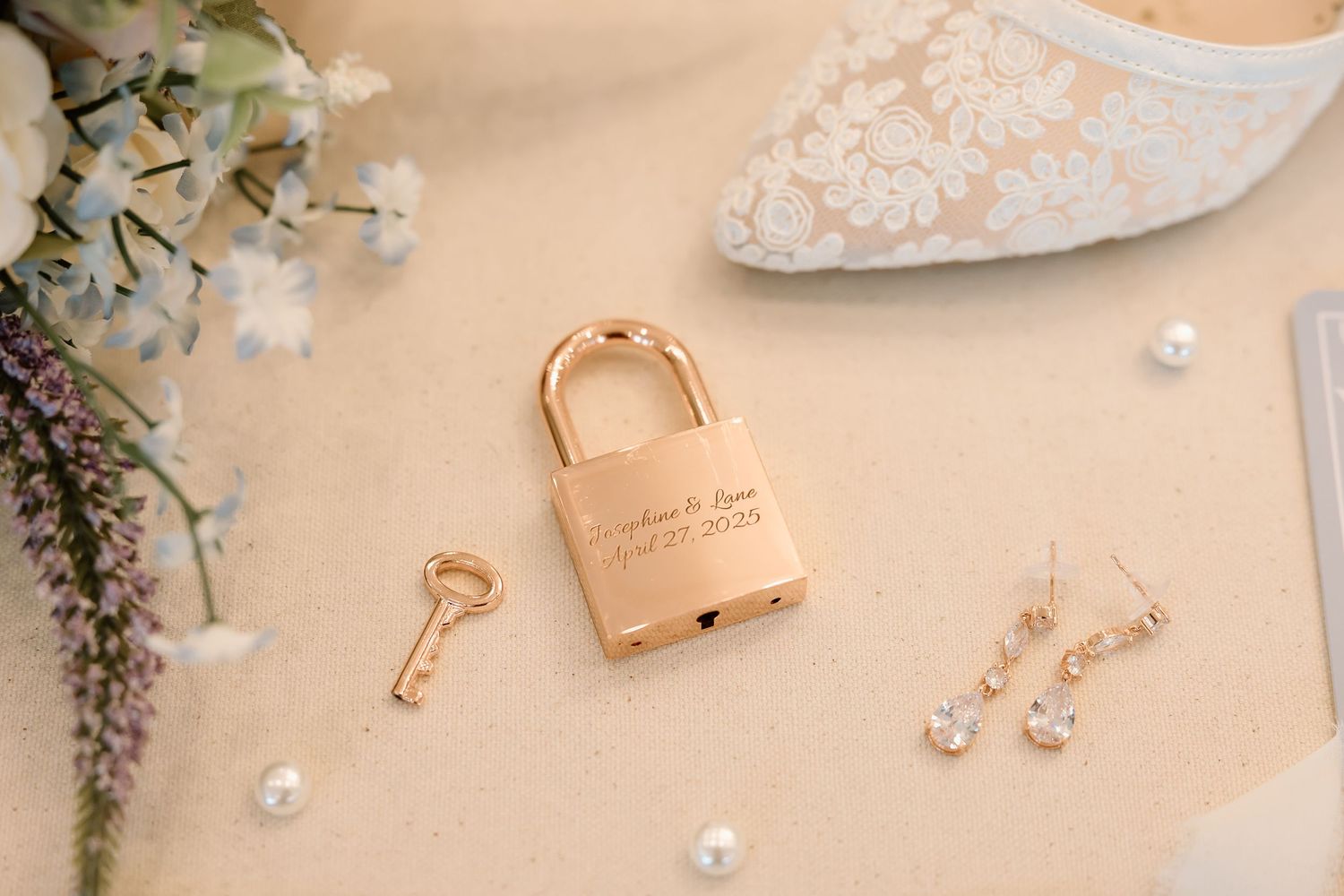 Wedding accessories including a gold love lock with key, delicate pearl earrings, and lace bridal shoe on beige background.