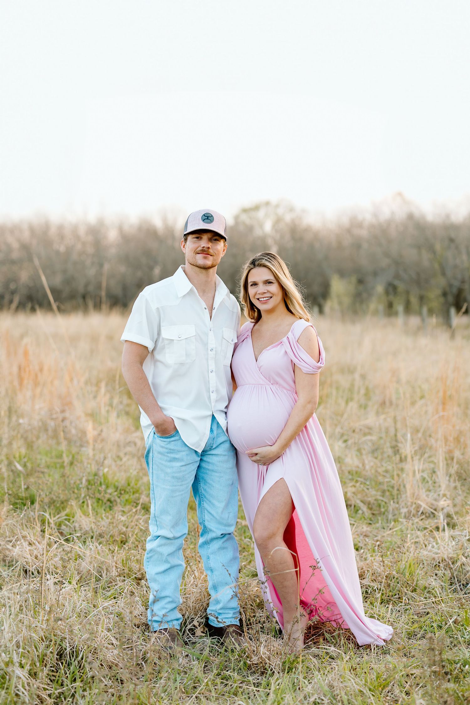 A couple stands together in a golden field, with the woman wearing a pink dress showing her pregnant belly.