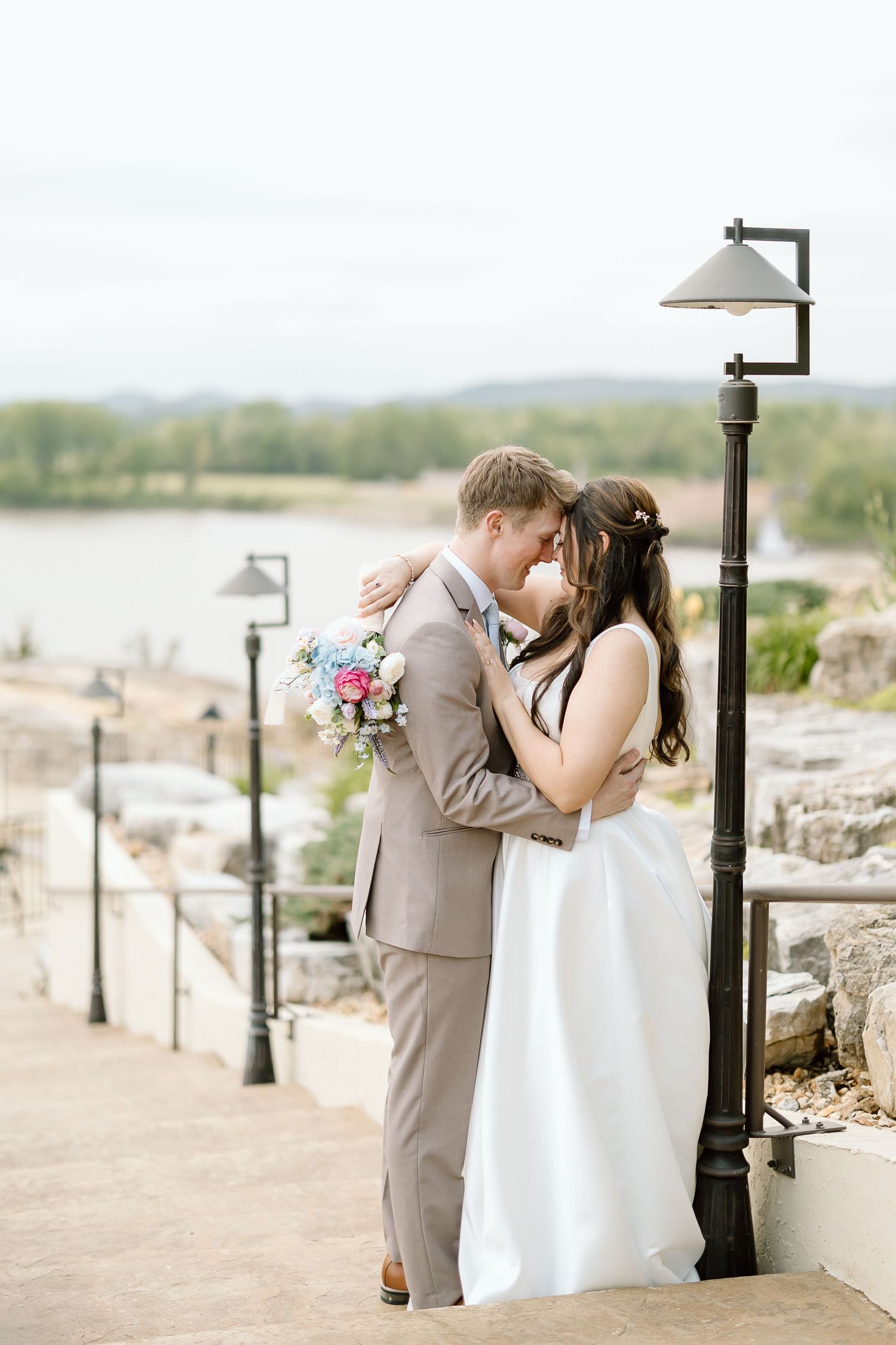 A romantic wedding couple embraces on stone steps by a lake with vintage lampposts and a pink floral bouquet.