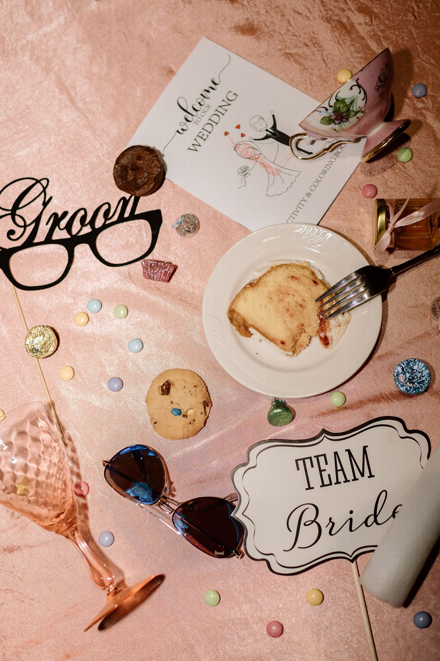 Wedding accessories including Team Bride sign, sunglasses, cookies, and invitation details arranged on pink background.