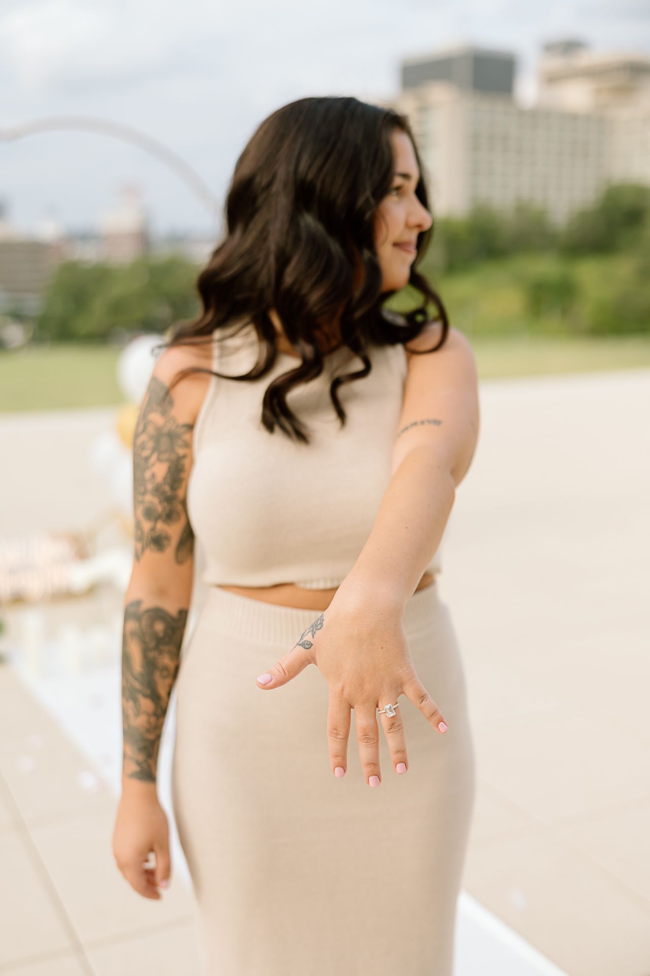 A person in a cream-colored dress and white crop top poses outdoors showing their arm tattoo and engagement ring.
