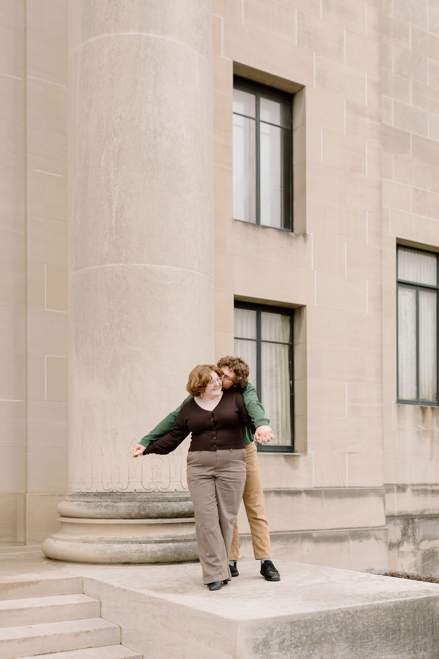 A couple sits and leans back on concrete steps in front of a stone building with windows.