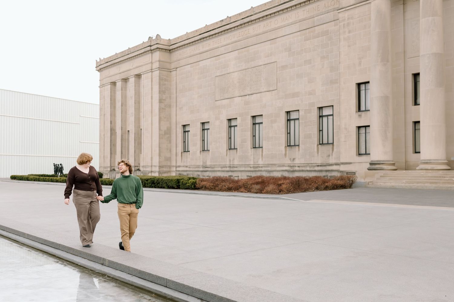 Two people walking together along a concrete path outside a large stone building with classical architecture.