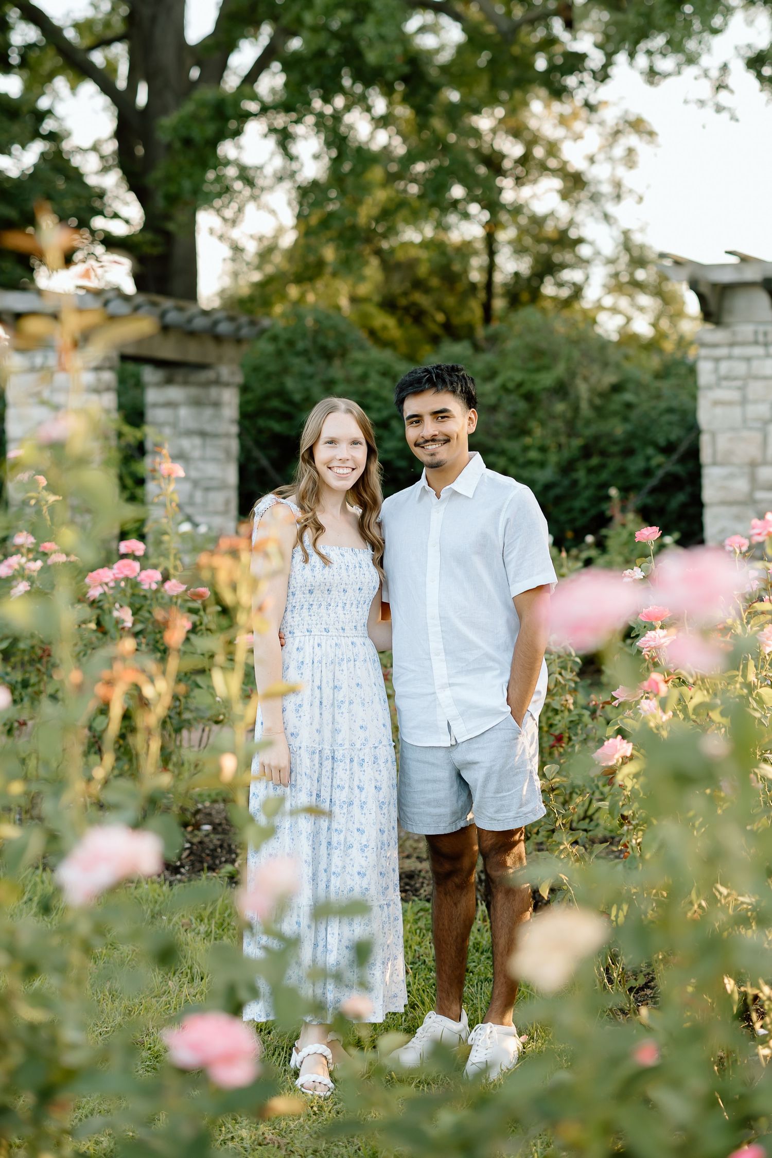 A couple stands together in a garden with pink flowers and stone pillars during golden hour.