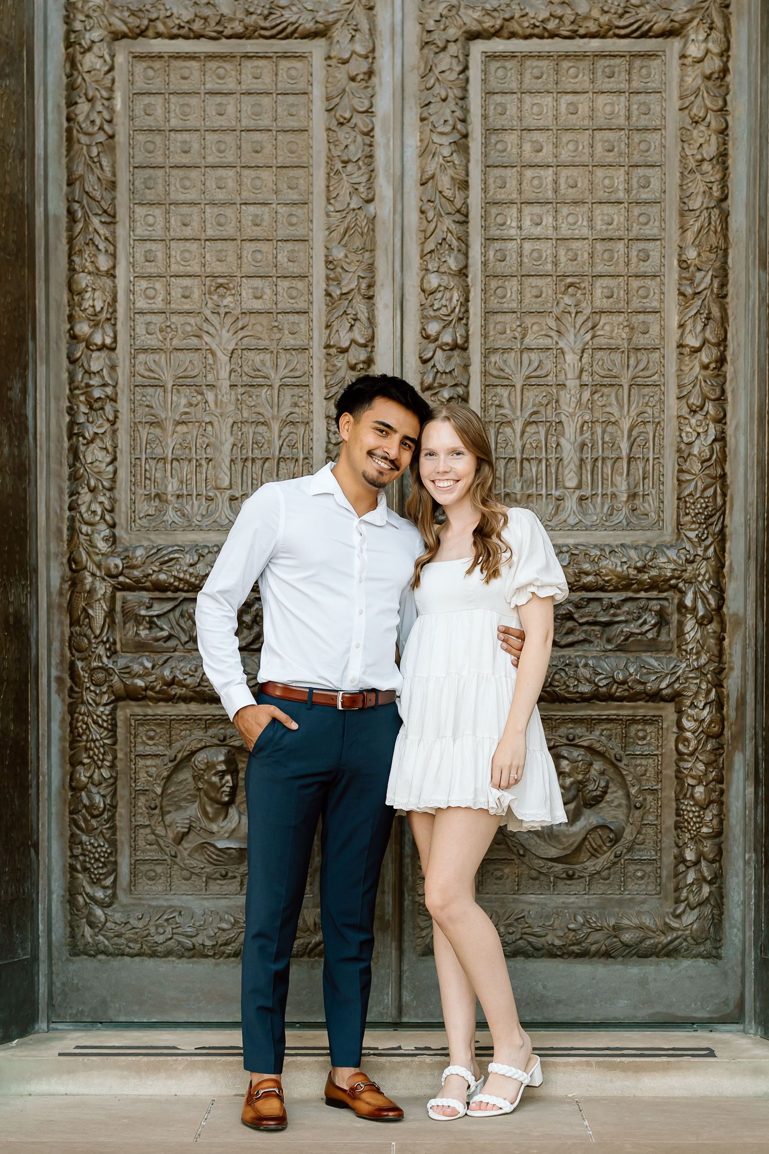 A couple poses playfully against an ornate carved stone wall in a romantic engagement photo shoot.