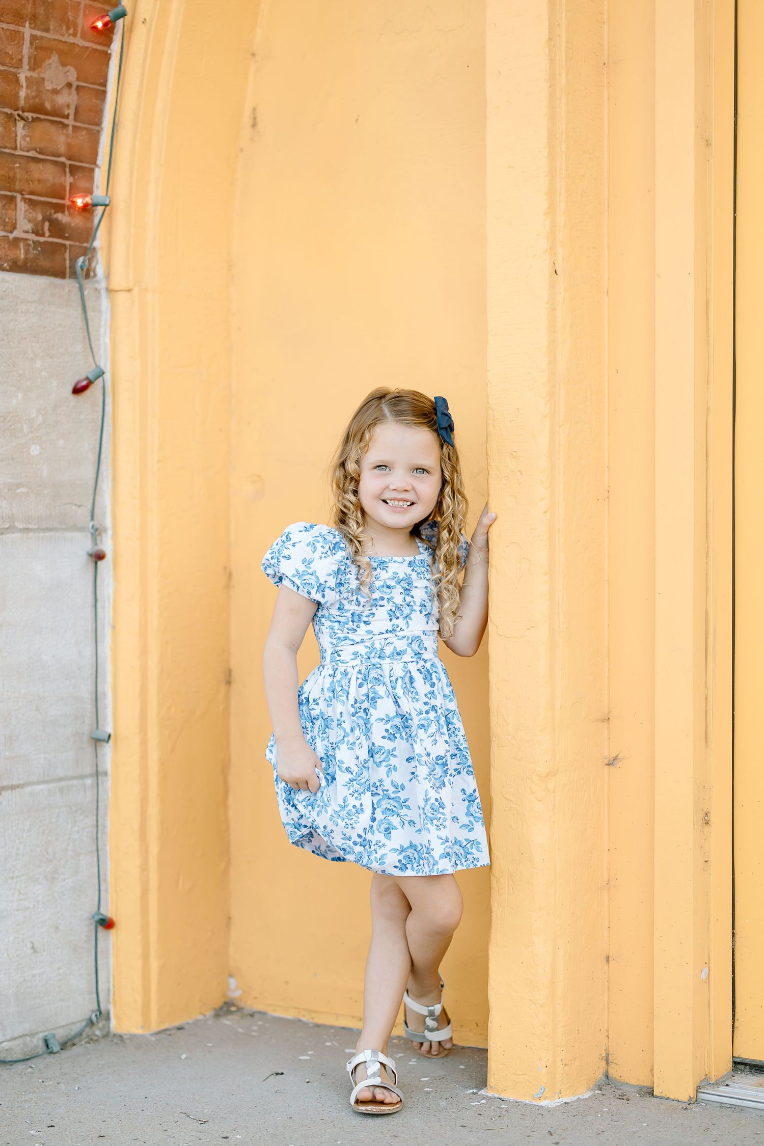 A child in a blue and white floral dress poses playfully against a yellow wall in a series of candid shots.