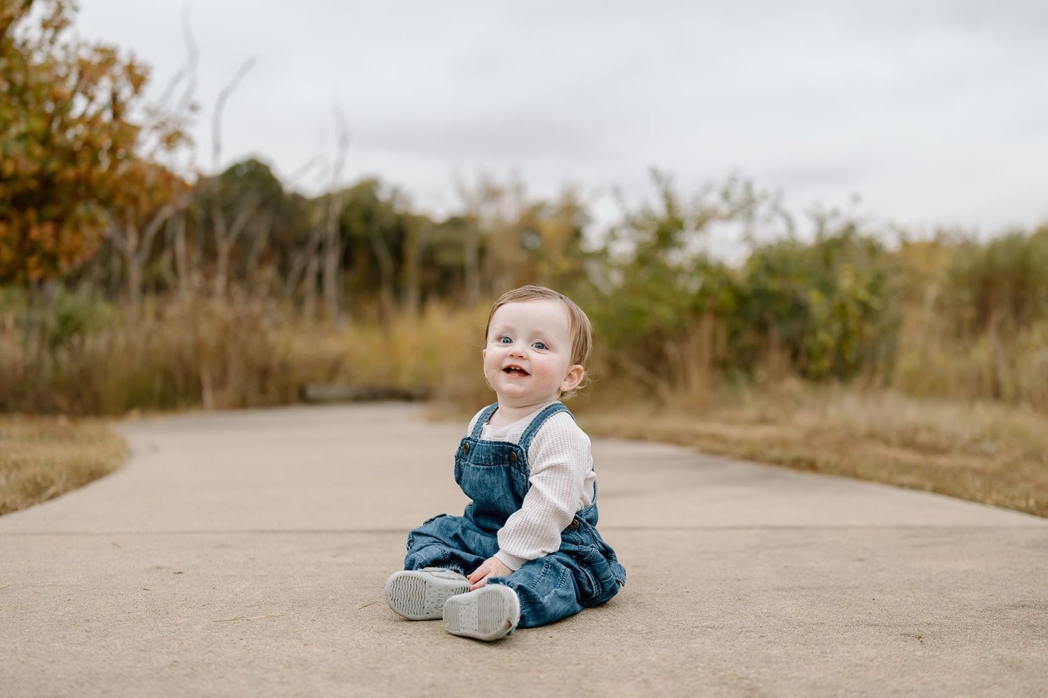 A child in blue overalls plays and lies down on a dirt path surrounded by autumn foliage.