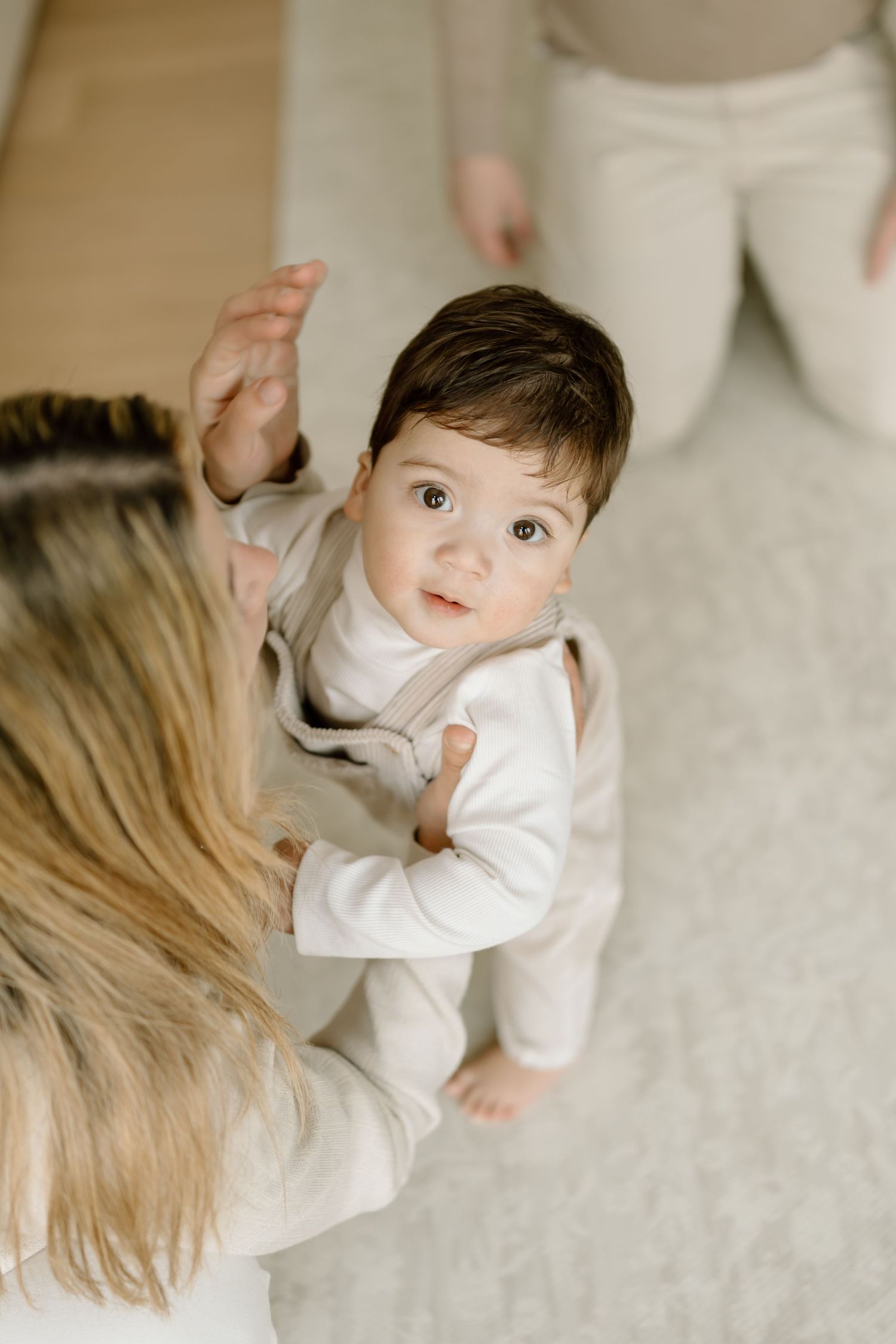 A small child in white clothing stands while reaching up towards someone on a light carpet.