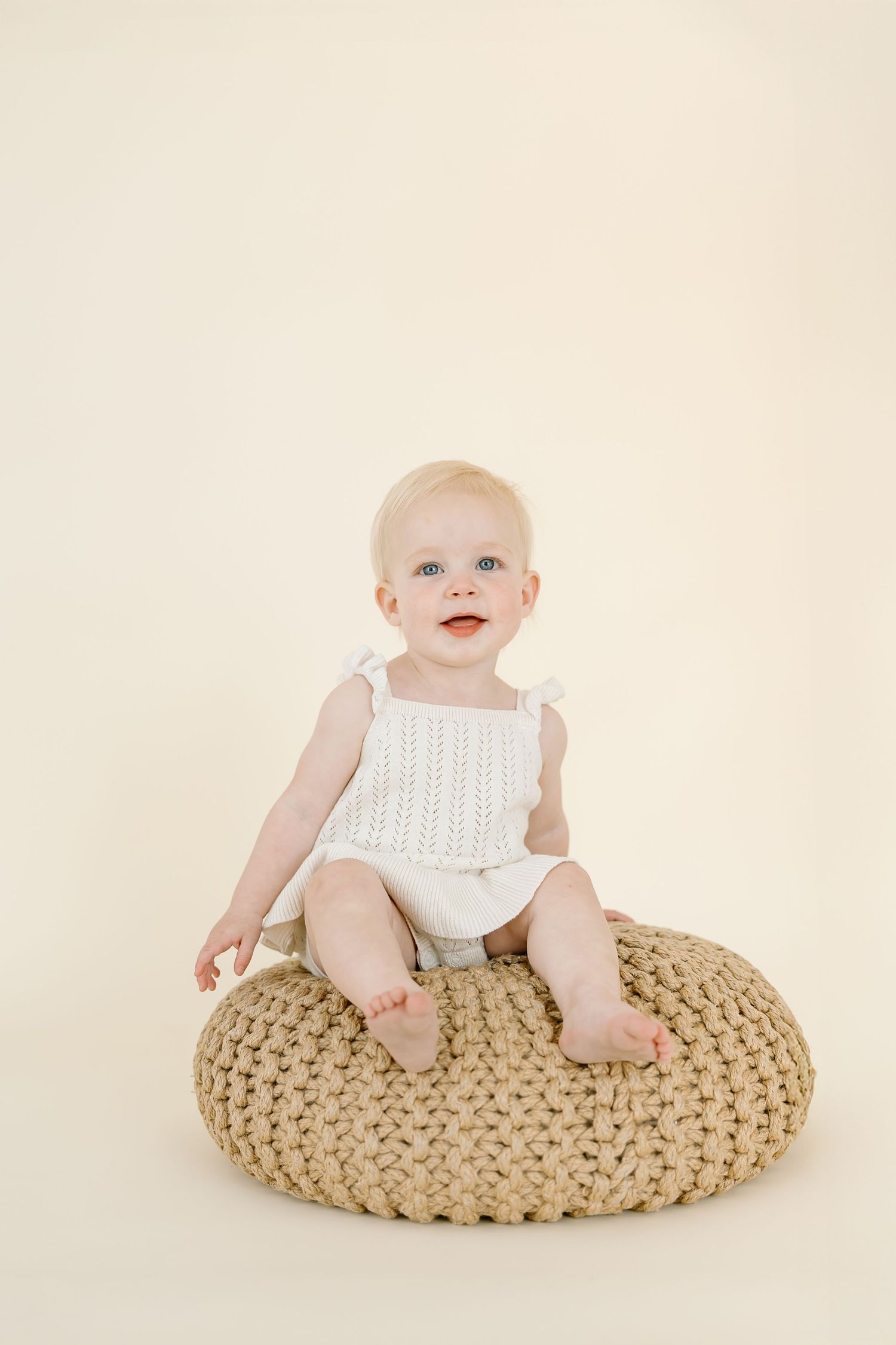 A baby wearing a white top sits and smiles on a large beige knitted pouf against a cream background.