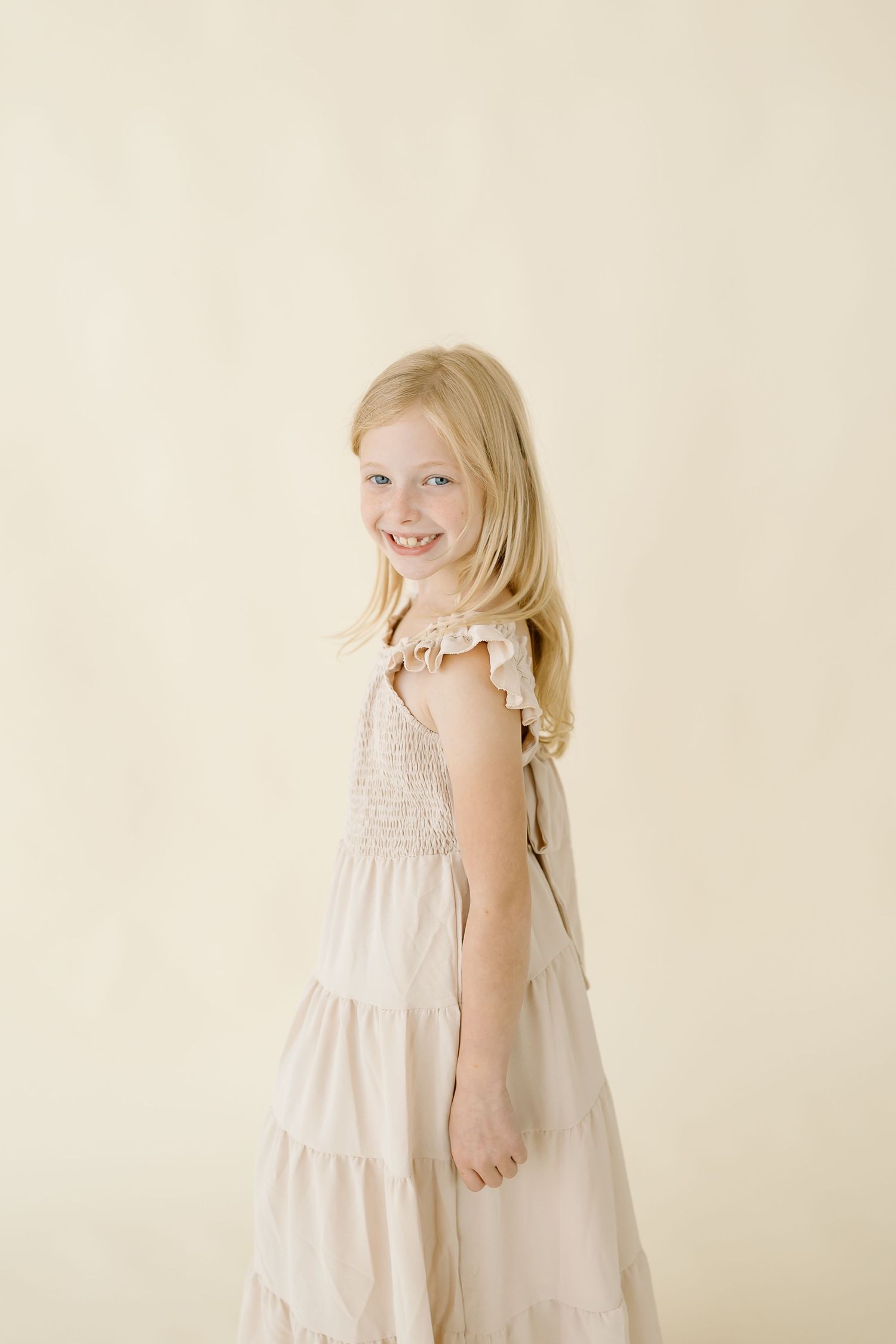 Young child in a flowing cream-colored tiered dress with ruffled sleeves poses against a light beige background.