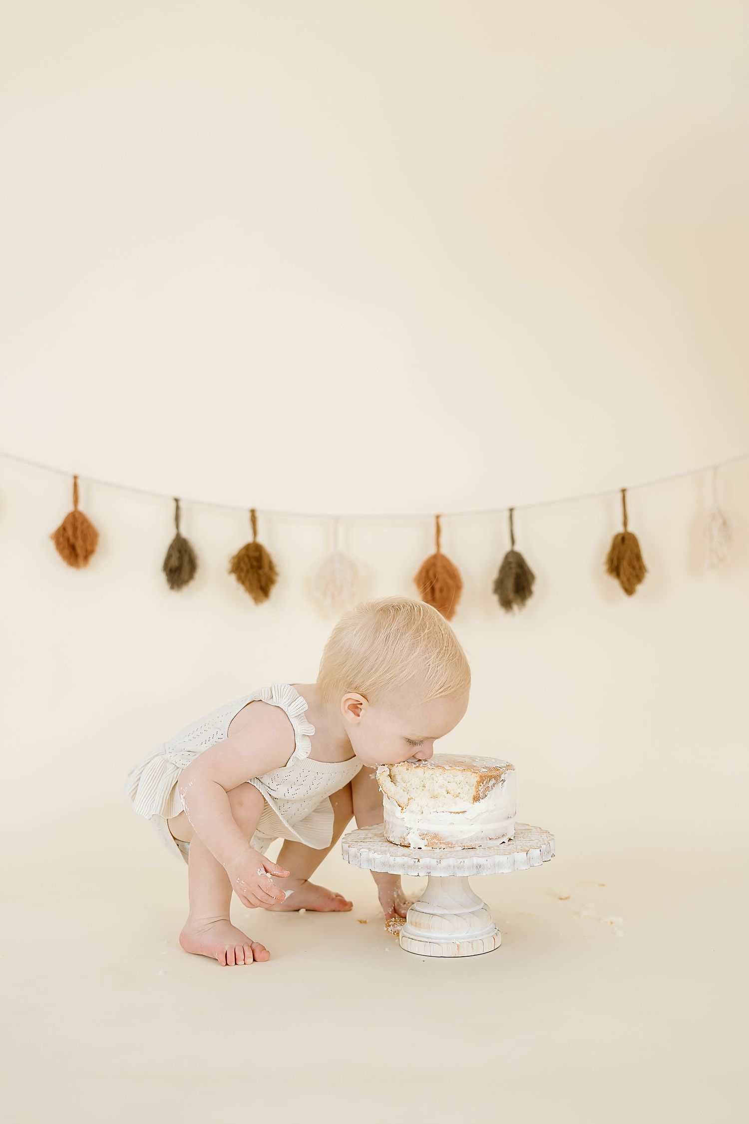 Baby in white outfit leans forward to taste a tiered white cake with autumn leaf garland hanging on wall behind.