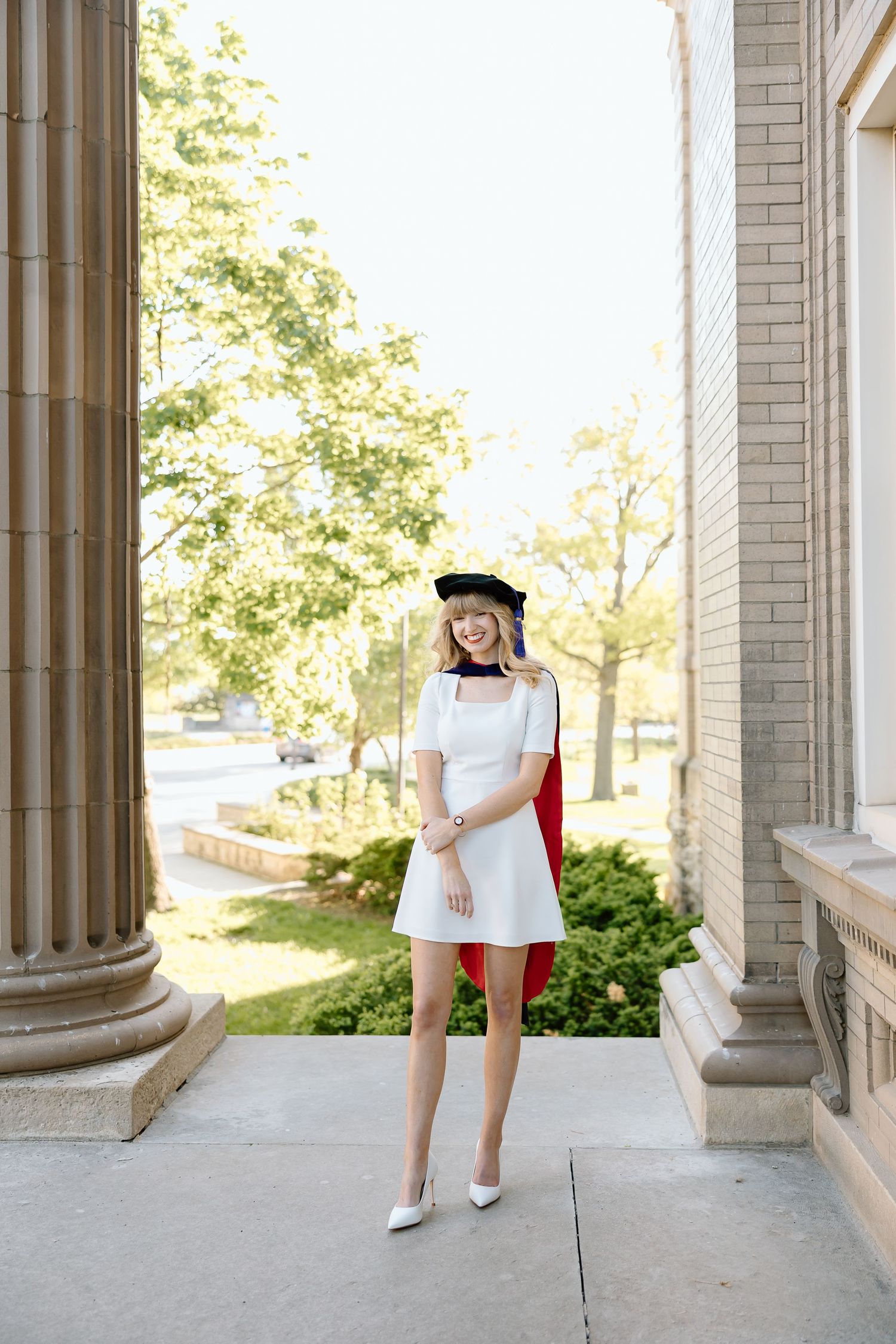 Graduate in red dress and graduation cap poses with diploma near stone columns on sunny day.