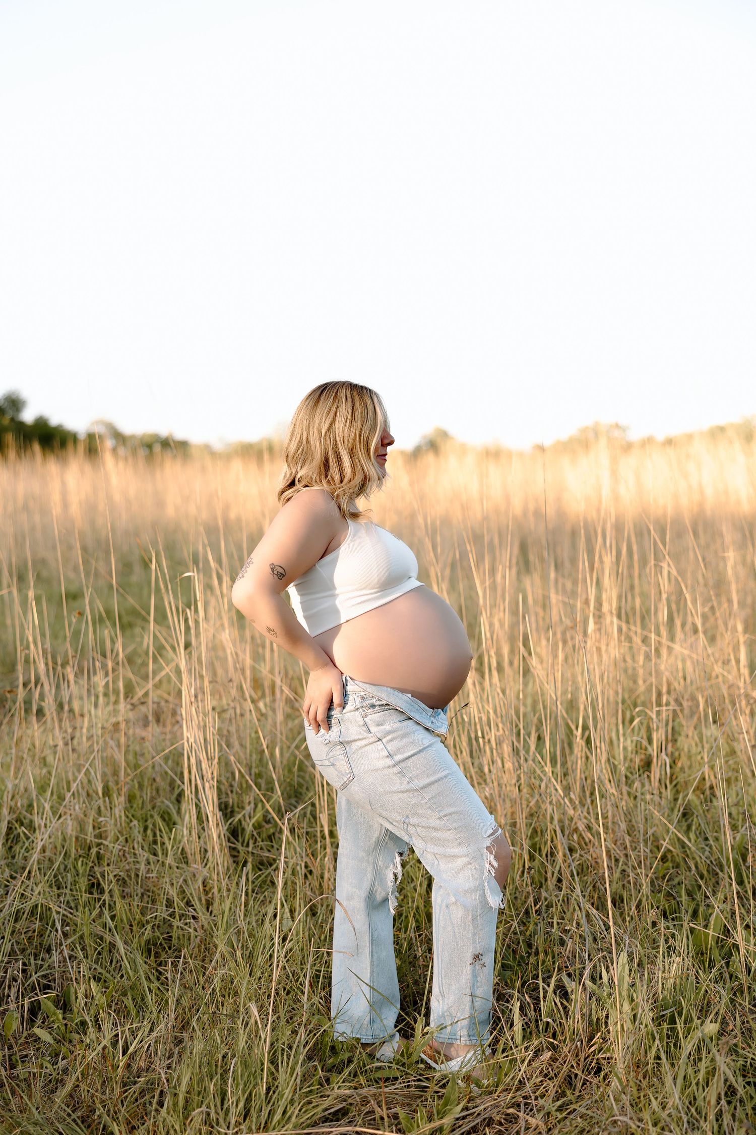 A pregnant woman in jeans and a white top stands in a golden field at sunset.