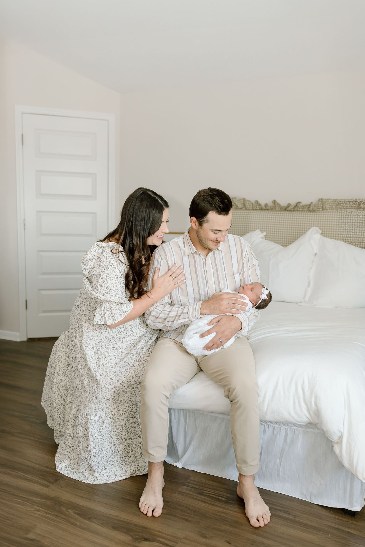 A couple sits on a white bed together sharing tender moments with their newborn baby in a bright bedroom.