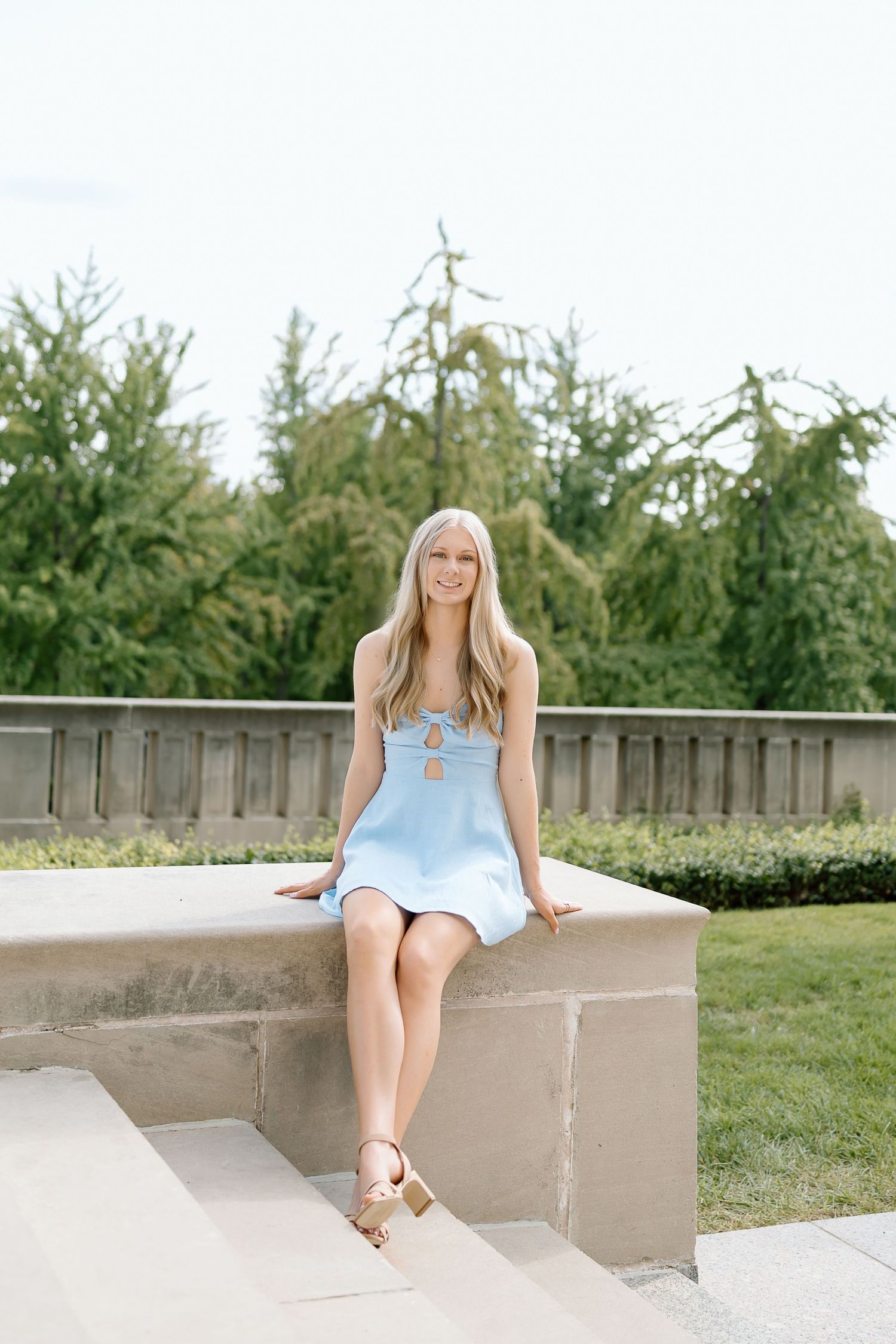 A person in a light blue dress sits casually on concrete steps in an outdoor setting with greenery in the background.