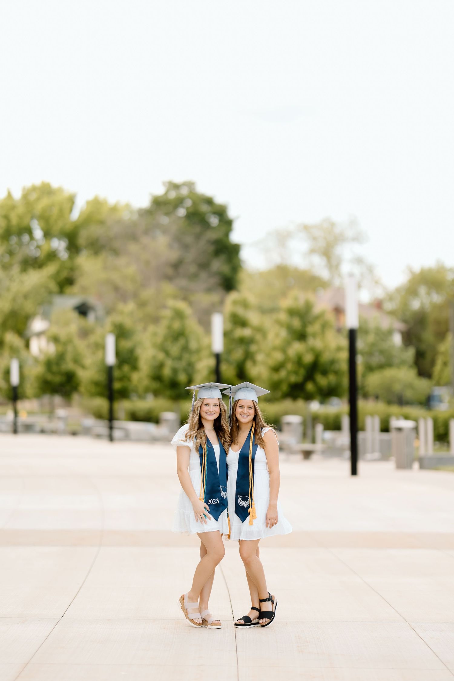 Two graduates in matching blue caps and gowns crouch together on concrete walkway.