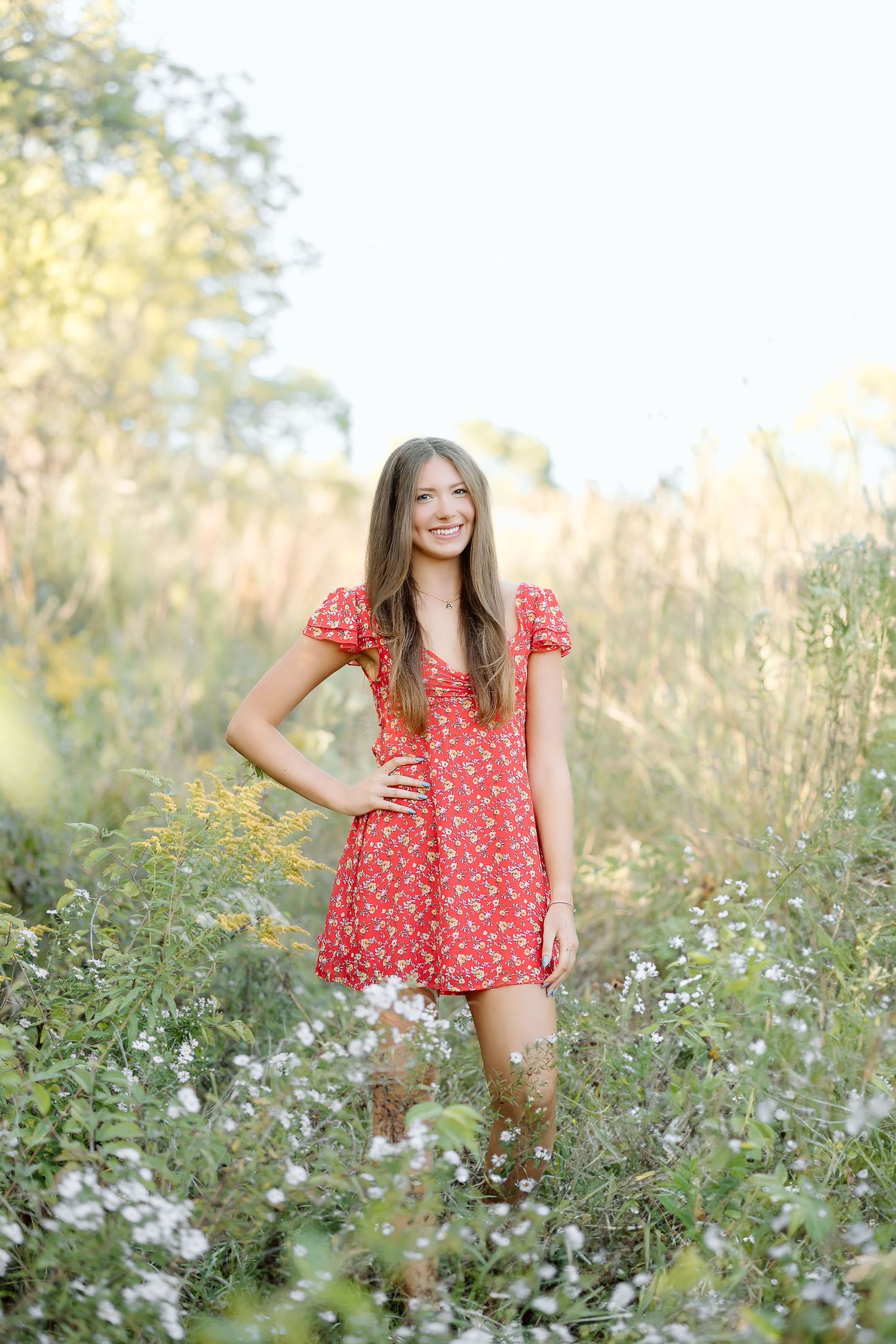 A person in a red floral dress poses joyfully among white wildflowers in a sunny meadow during golden hour.