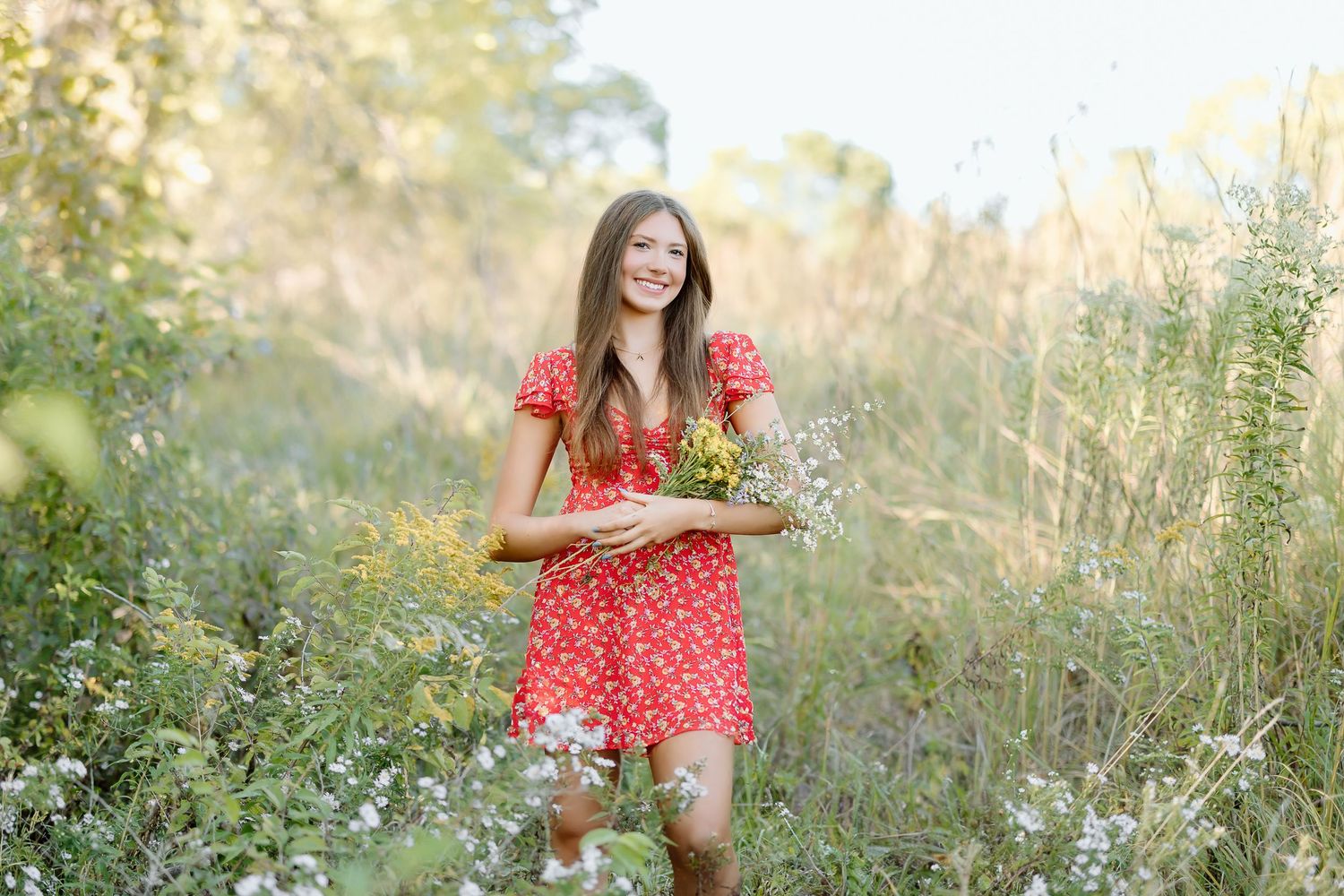A person in a red polka dot dress poses in a sunlit meadow with tall grass and wildflowers during golden hour.