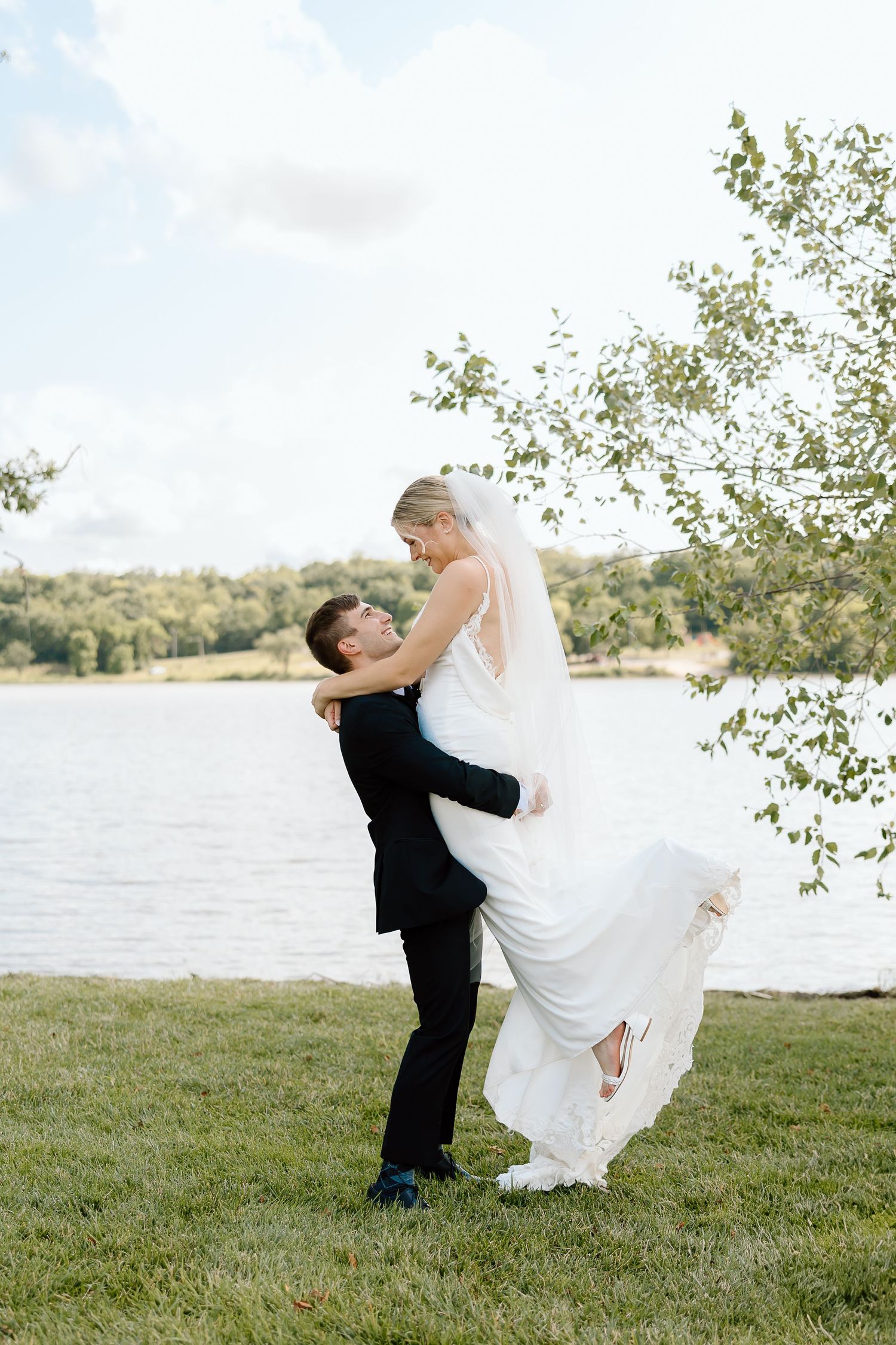 A couple in wedding attire performs playful dance moves by a scenic lakeside setting on a grassy shore.