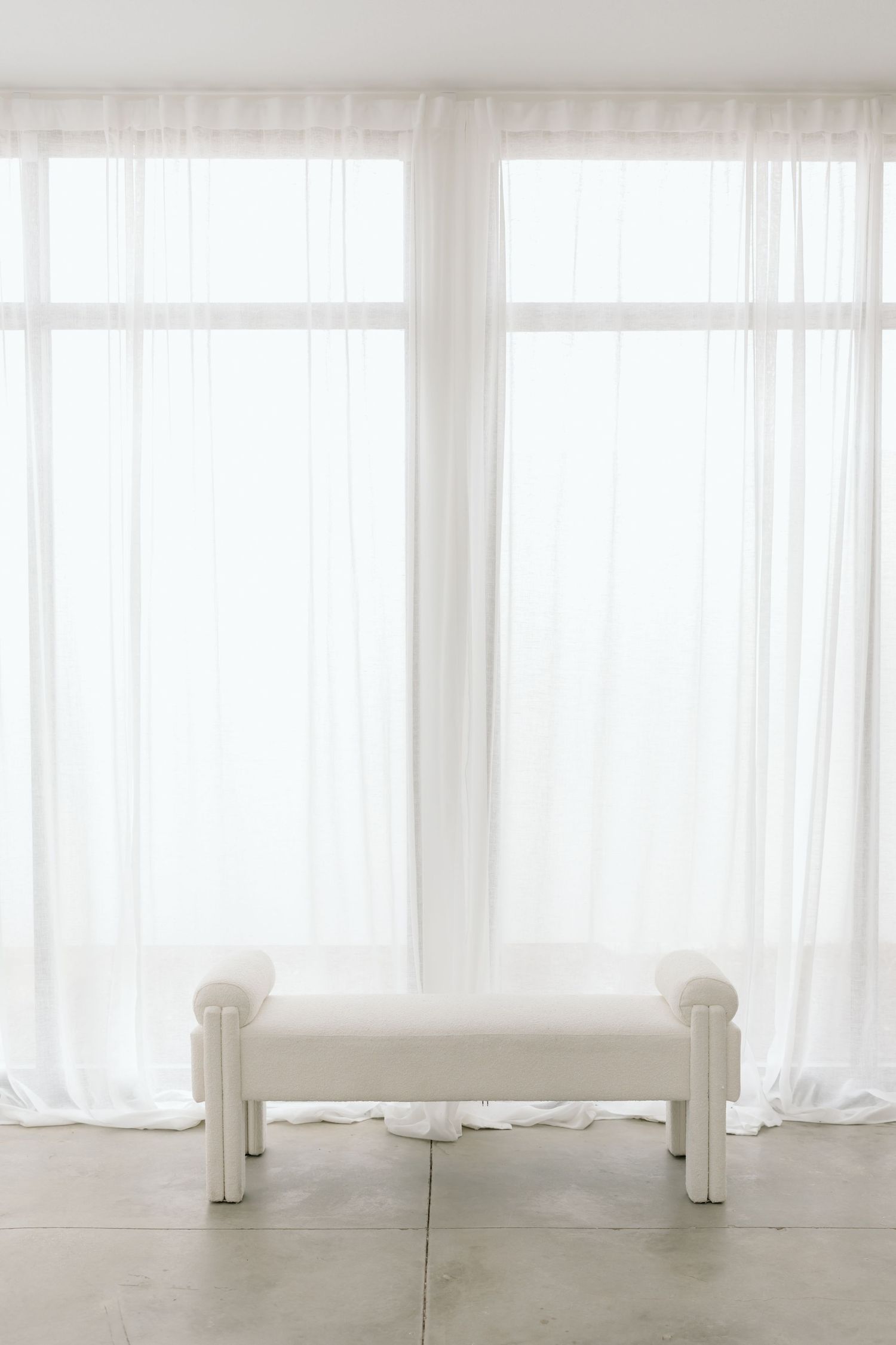 White sheer curtains hanging in front of bright windows create a soft, ethereal atmosphere in a minimalist interior space.
